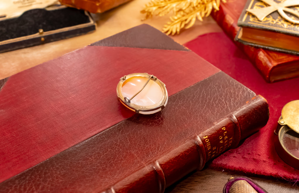 A vintage-style round pendant with a pale pinkish-white stone and gold-toned setting, resting on a dark red leather-bound book surrounded by antique books and props.
