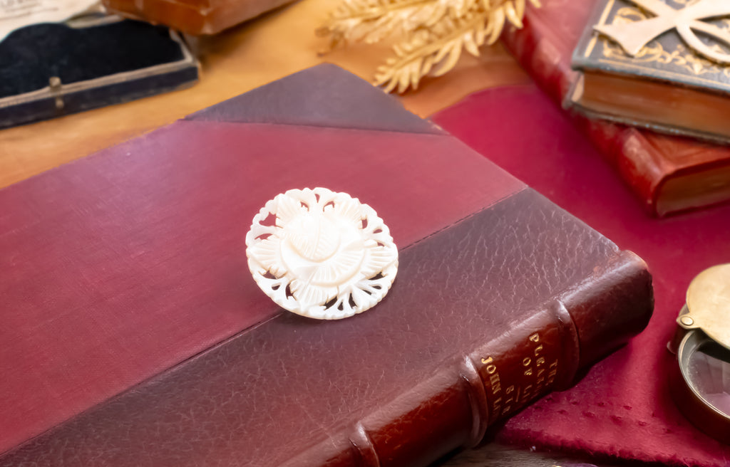 A round white carved mother of pearl brooch with a floral pattern, displayed on top of a dark reddish-brown leather-bound book on a wooden desk surrounded by antique books.