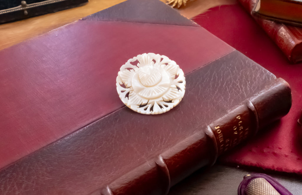 A close-up of a carved mother of pearl  circular brooch with a floral sunburst design, resting on the cover of a dark reddish-brown leather-bound book.