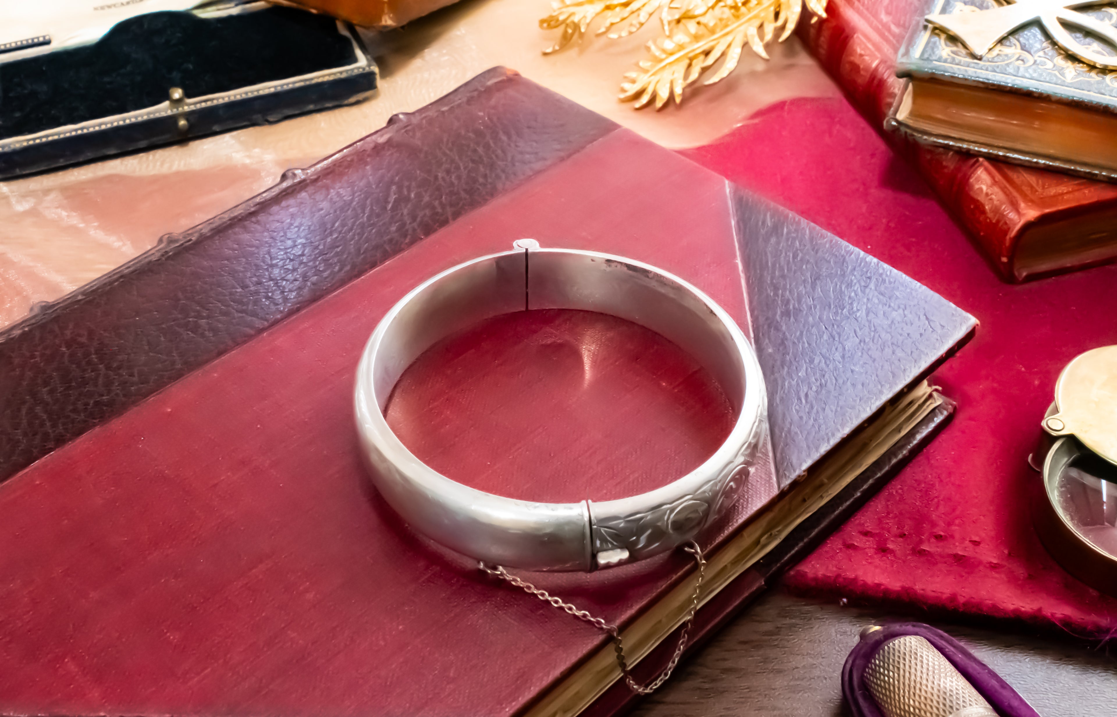 A polished silver bangle bracelet with engraved details and a small attached chain, displayed on a dark red leather book surrounded by vintage books and accessories.