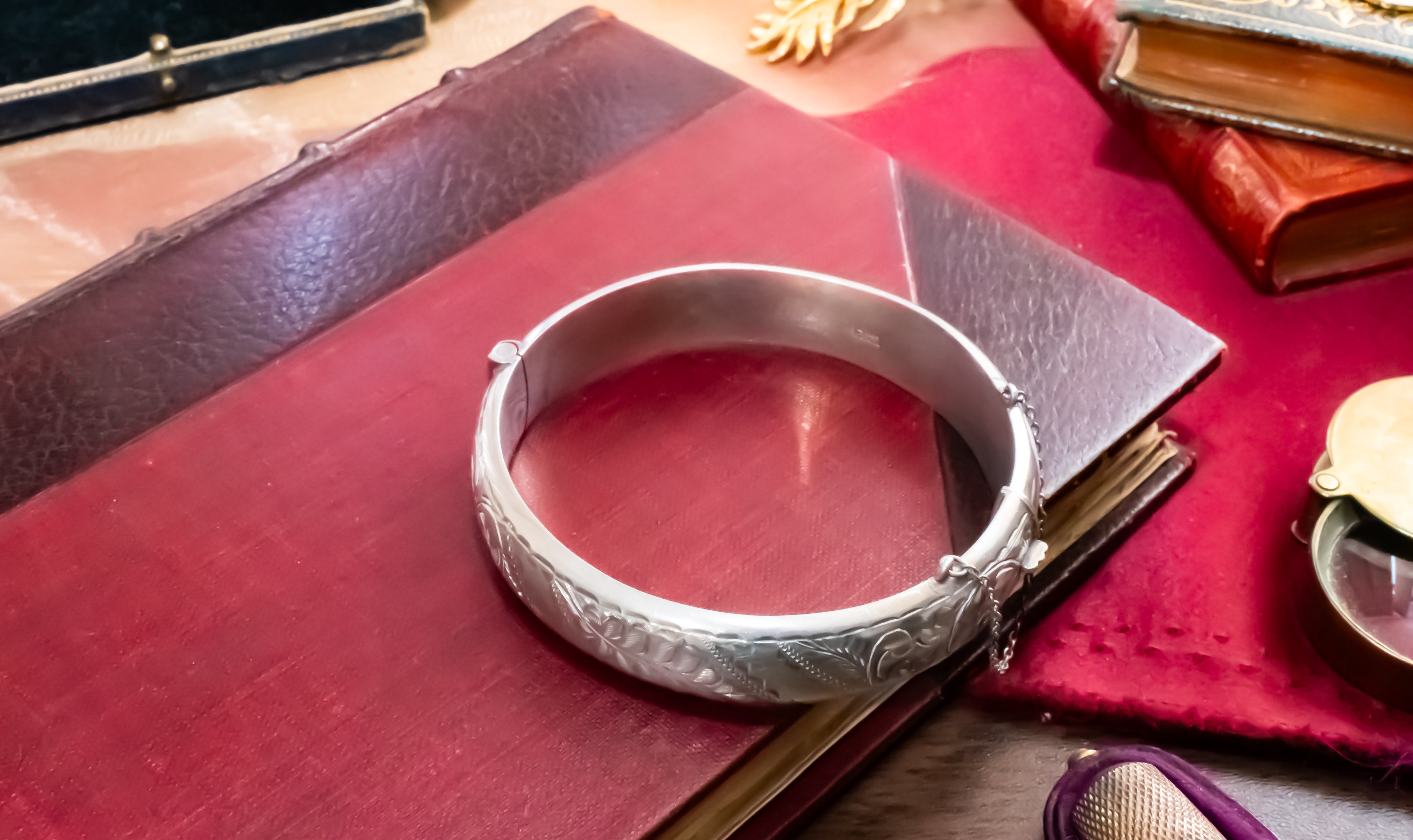A polished silver-toned bangle bracelet with engraved floral patterns, resting on a dark red leather journal surrounded by vintage books and a red cloth.