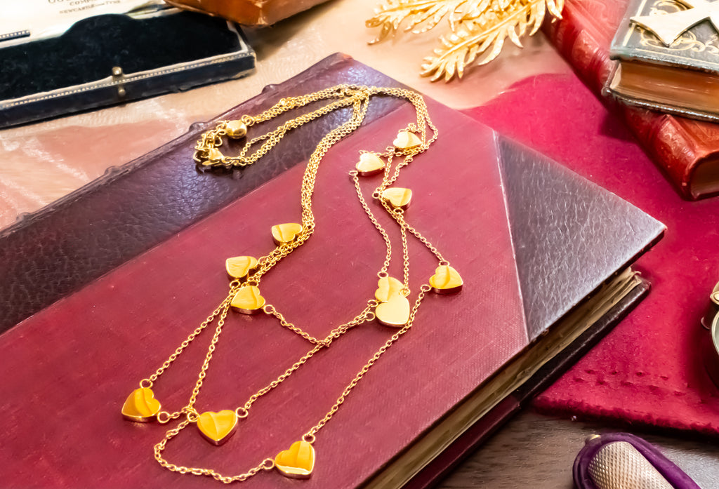 A gold heart-link necklace with multiple small heart charms draped over a dark red book, surrounded by vintage-style props on a wooden surface.