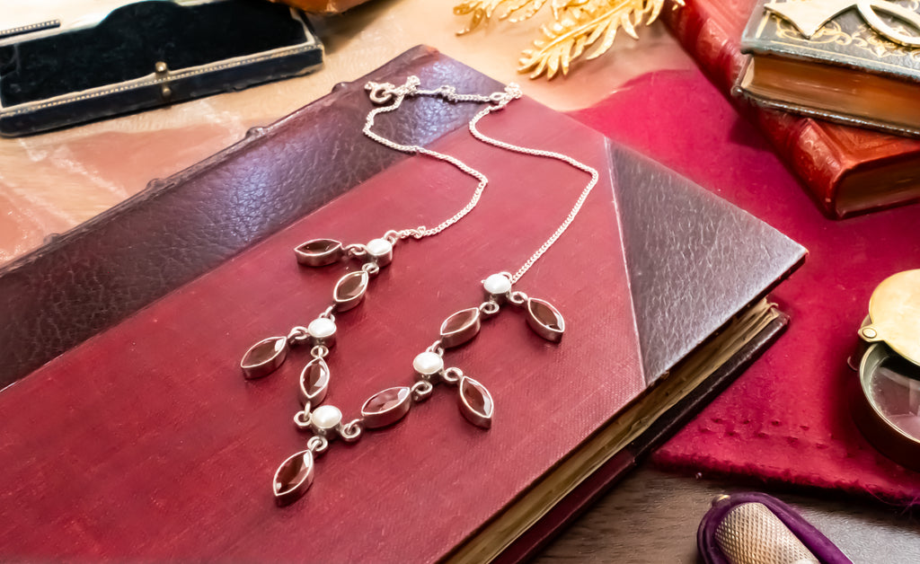 A silver-toned necklace with marquise-shaped dark brown stones and small white pearl accents, displayed on an antique red leather book with vintage props around it.