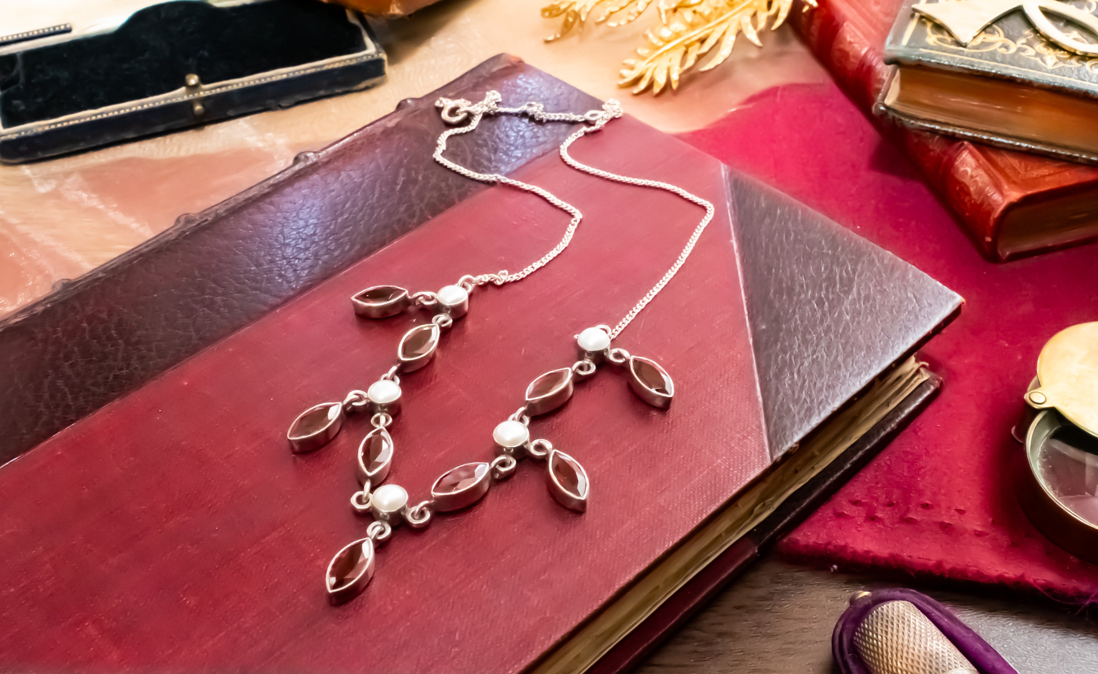 A silver-toned necklace with marquise-shaped dark brown stones and small white pearl accents, displayed on an antique red leather book with vintage props around it.