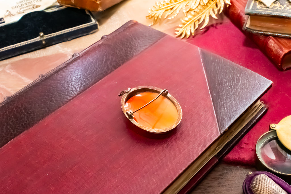 An oval gold-toned pin with a clear faceted gemstone and a safety pin clasp, resting on a dark red leather-bound book surrounded by vintage props.