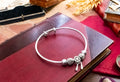 A silver bangle bracelet with decorative filigree beads and dangling charms, displayed on a dark red leather book with vintage props around it.