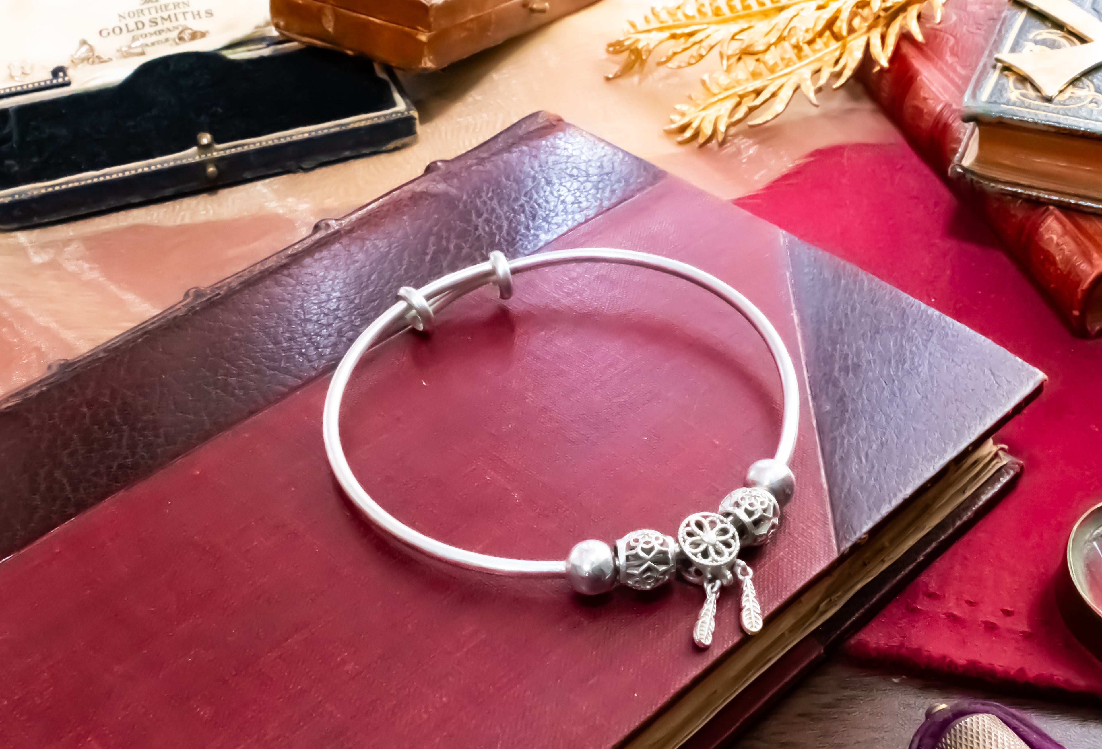 A silver bangle bracelet with decorative filigree beads and dangling charms, displayed on a dark red leather book with vintage props around it.