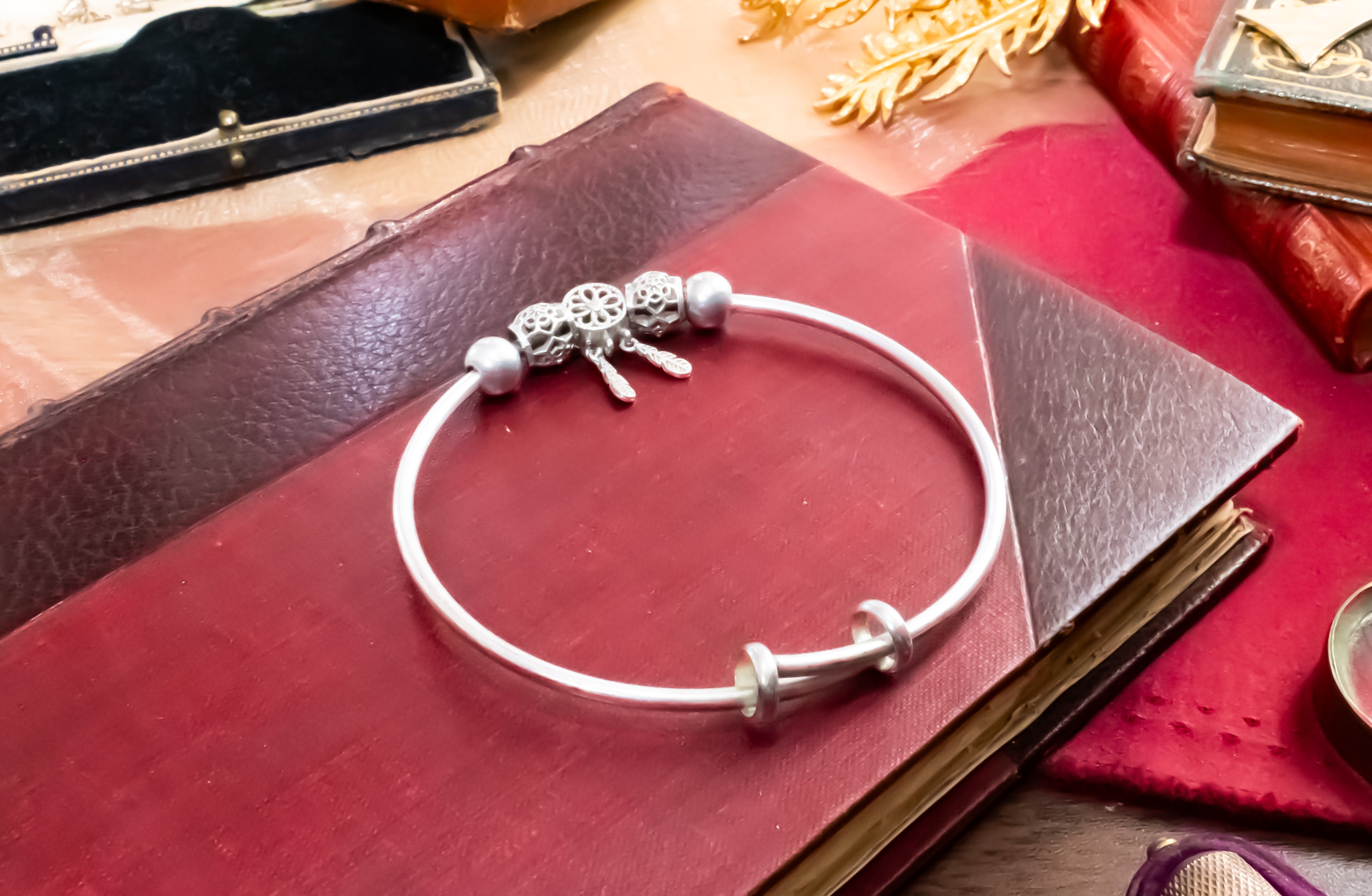 A silver-toned bangle bracelet with decorative charms, resting on a dark red leather book.