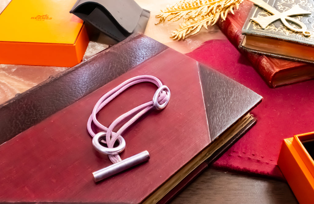 A light pink corded bracelet with a silver toggle clasp, displayed on a dark red leather journal surrounded by decorative books, an orange box, and a gold leaf ornament.