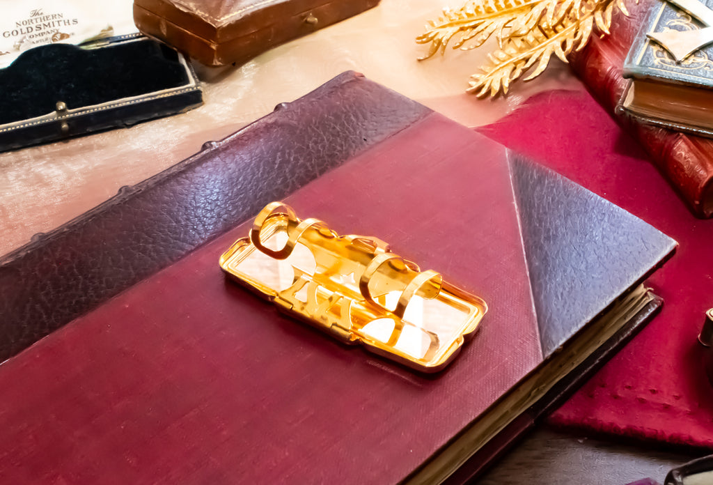 A gold-toned rectangular Stratton lipstick holder with a hinged mirror, resting on a dark red leather-bound book surrounded by vintage books and accessories.