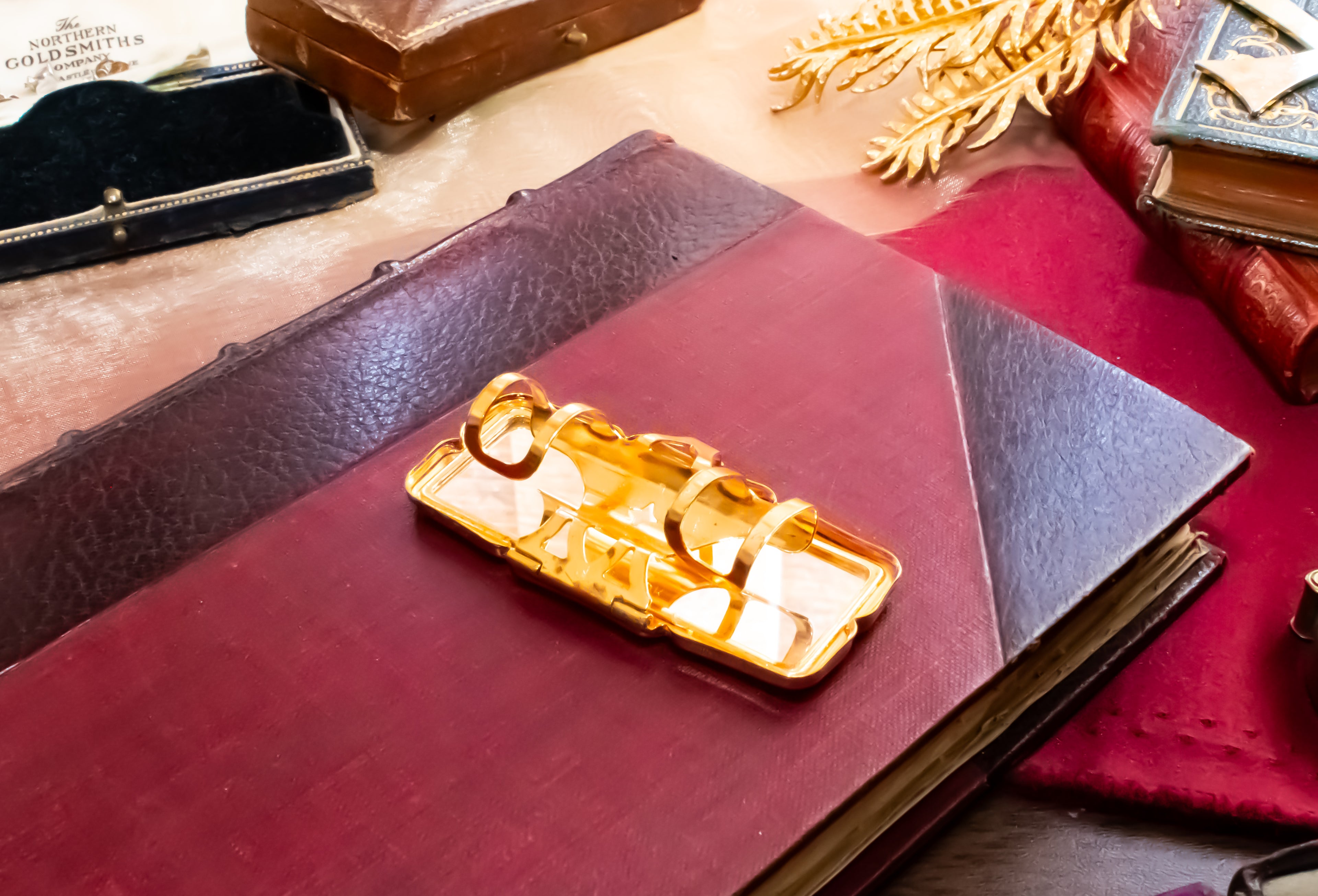 A gold-toned rectangular Stratton lipstick holder with a hinged mirror, resting on a dark red leather-bound book surrounded by vintage books and accessories.