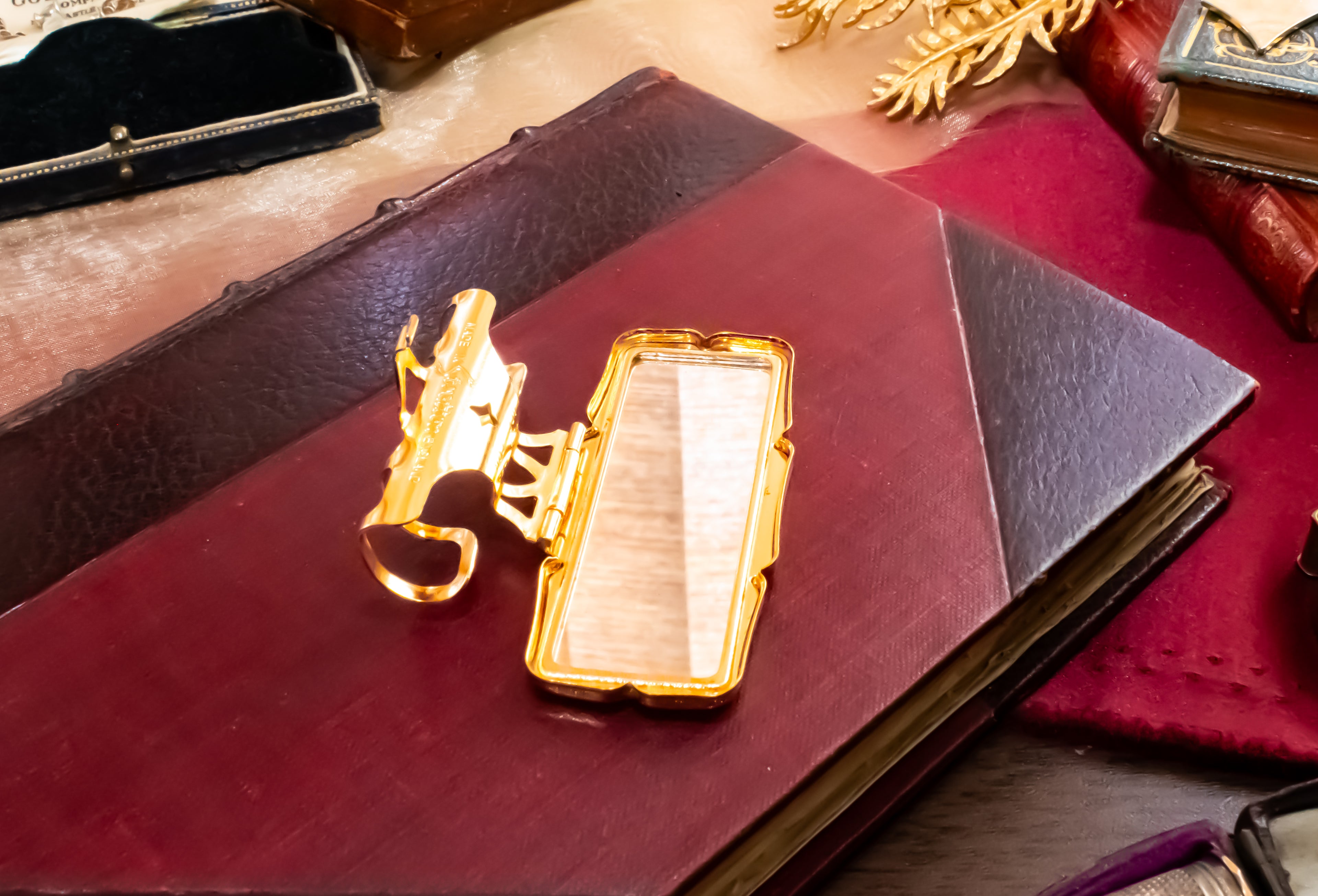 A gold-toned Stratton lipstick holder with a rectangular mirror face lies open on top of a dark red leather-bound book, surrounded by vintage props and fabric.