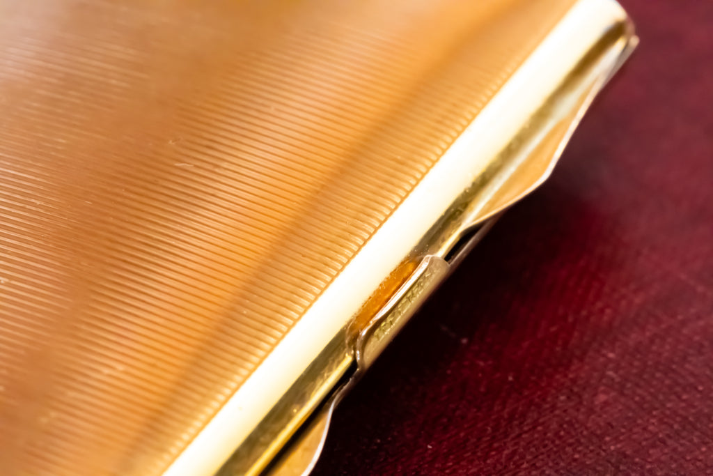 Close-up of a gold-toned metal Stratton powder compact with a ribbed textured surface, resting on a dark red fabric background.