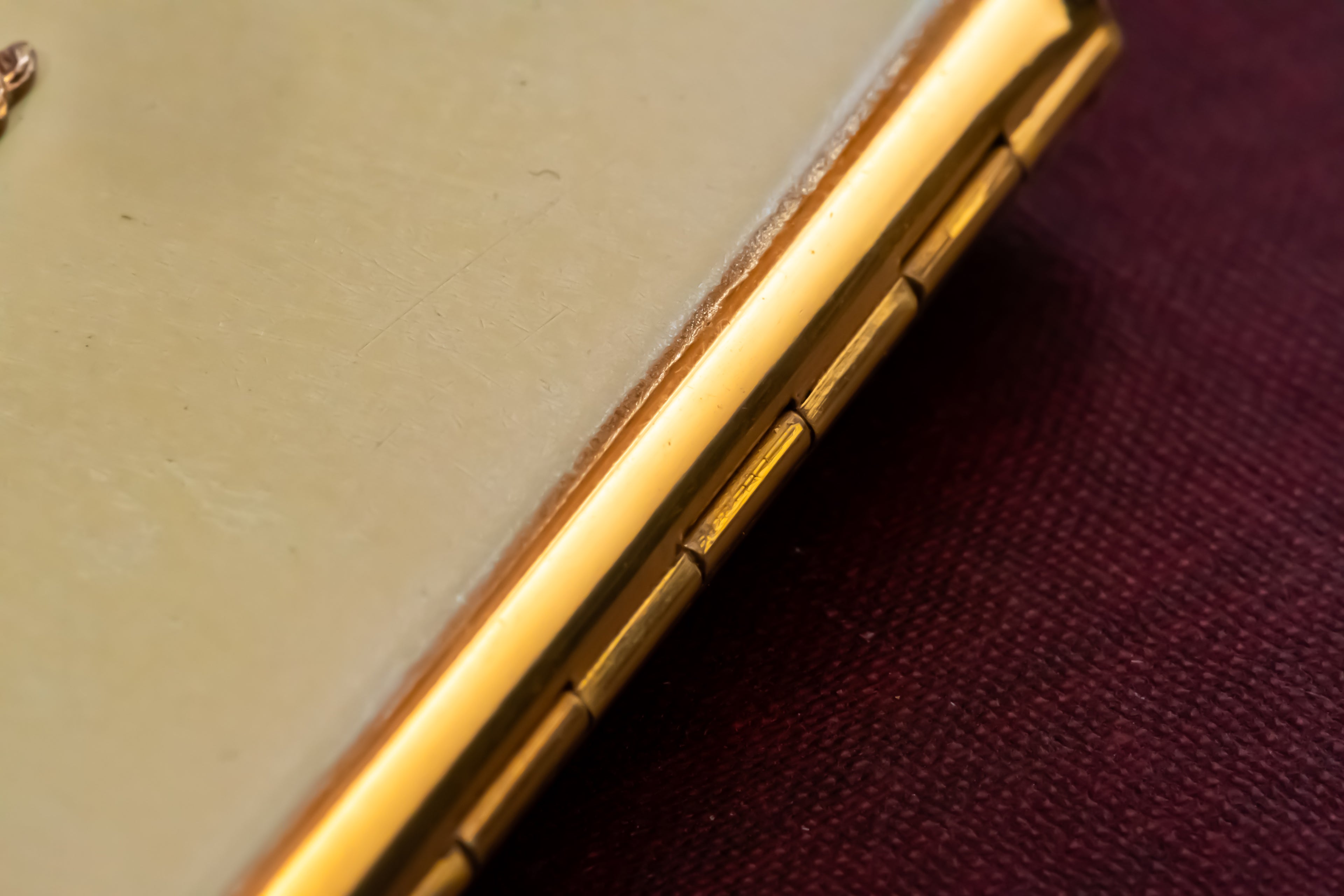 Close-up of a gold-toned metallic hinge or trim on a Stratton powder compact, set against a dark burgundy textured fabric background.