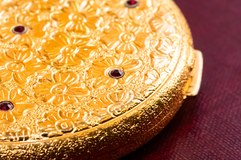 Close-up of a round gold compact or case with embossed floral detailing and small dark red jewel accents, resting on a dark burgundy surface.