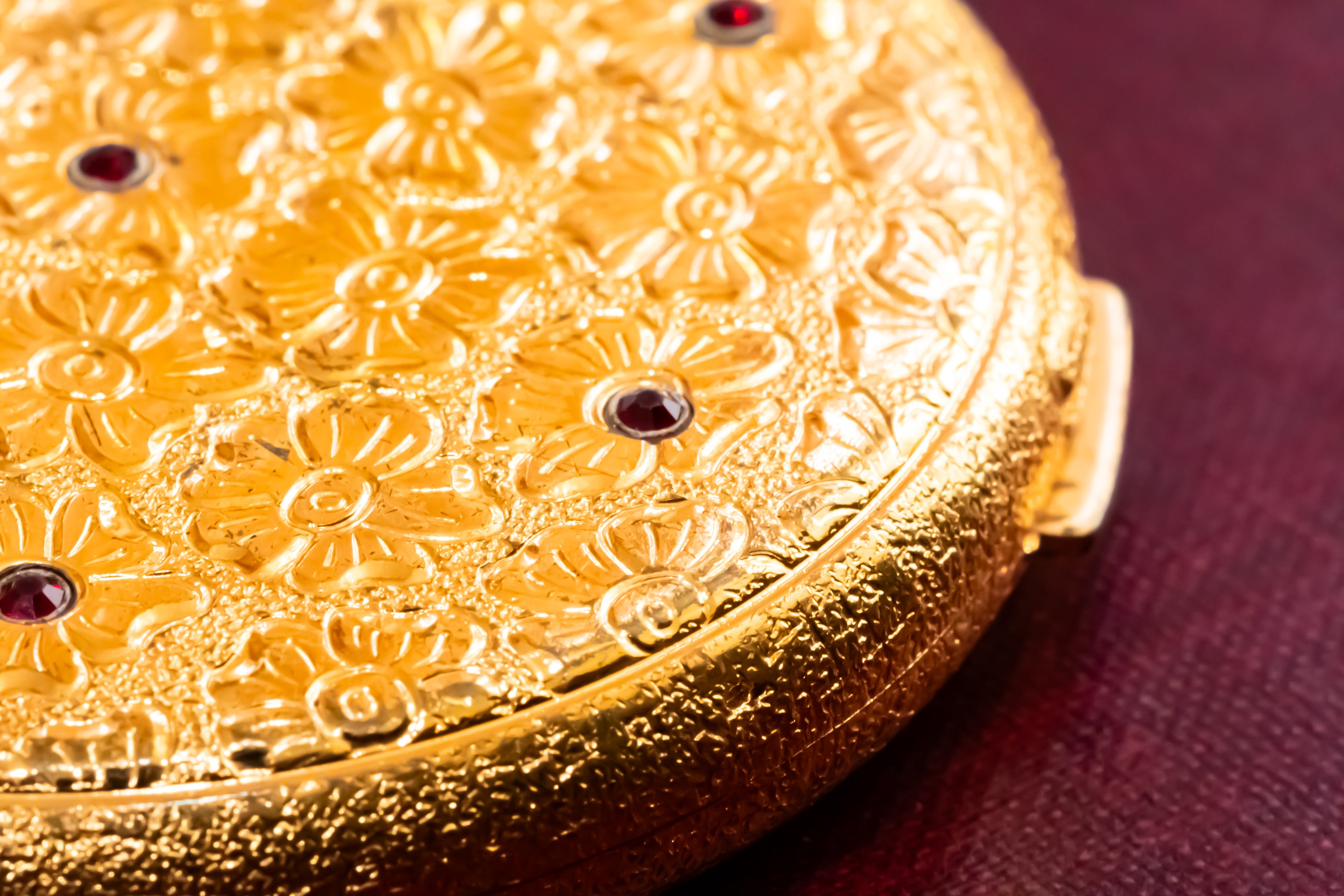 Close-up of a round gold compact or case with embossed floral detailing and small dark red jewel accents, resting on a dark burgundy surface.