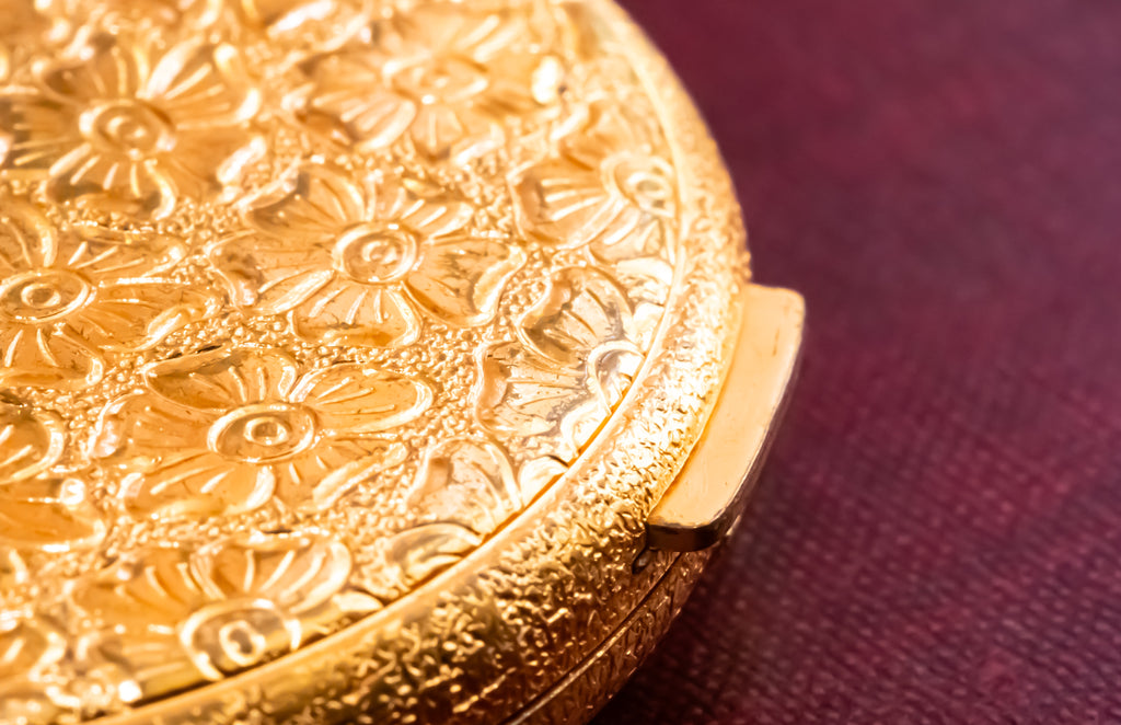 A close-up of an ornate gold-toned round compact or case with embossed floral detailing, resting on a dark burgundy textured surface.