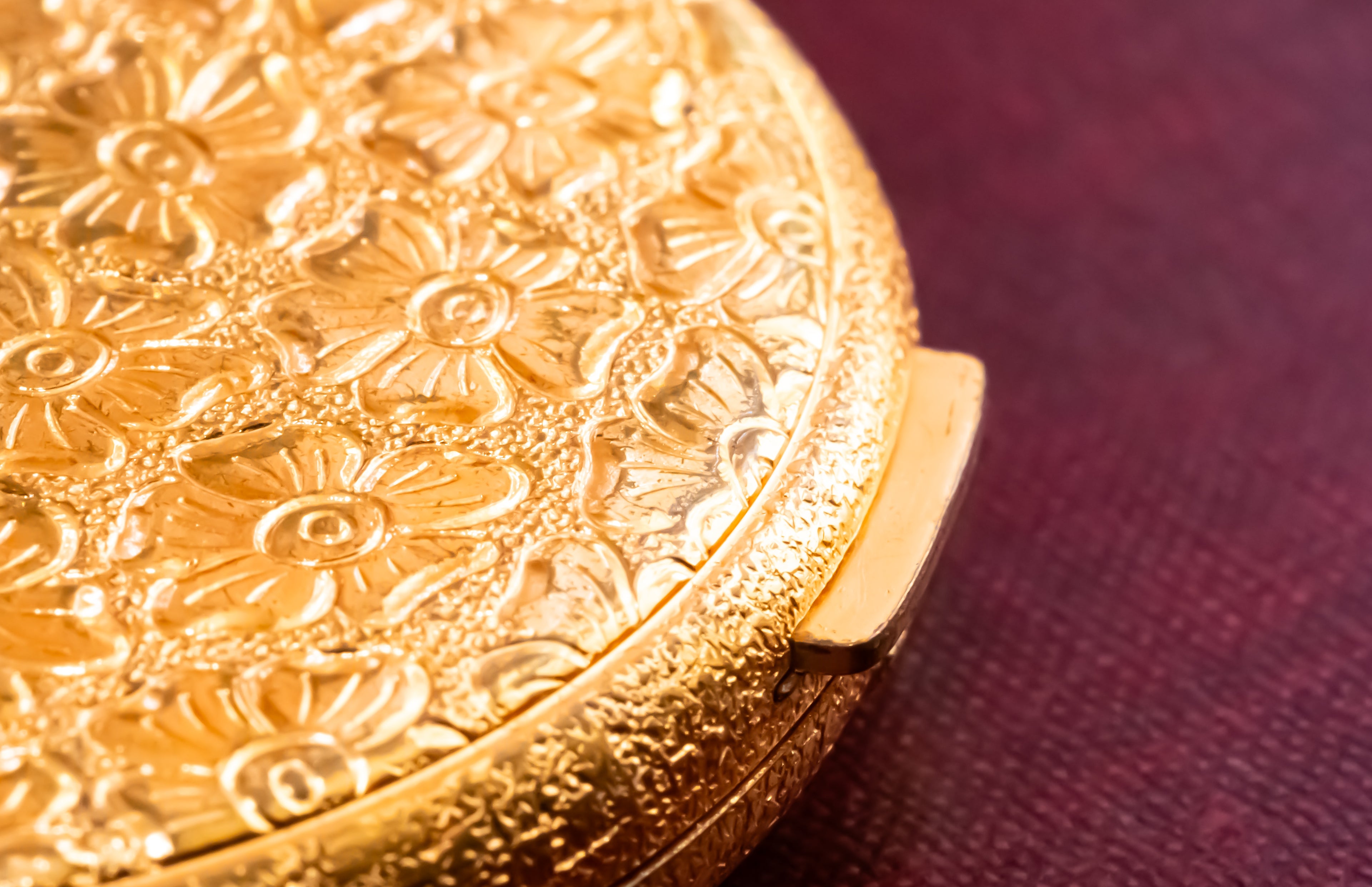 A close-up of an ornate gold-toned round compact or case with embossed floral detailing, resting on a dark burgundy textured surface.