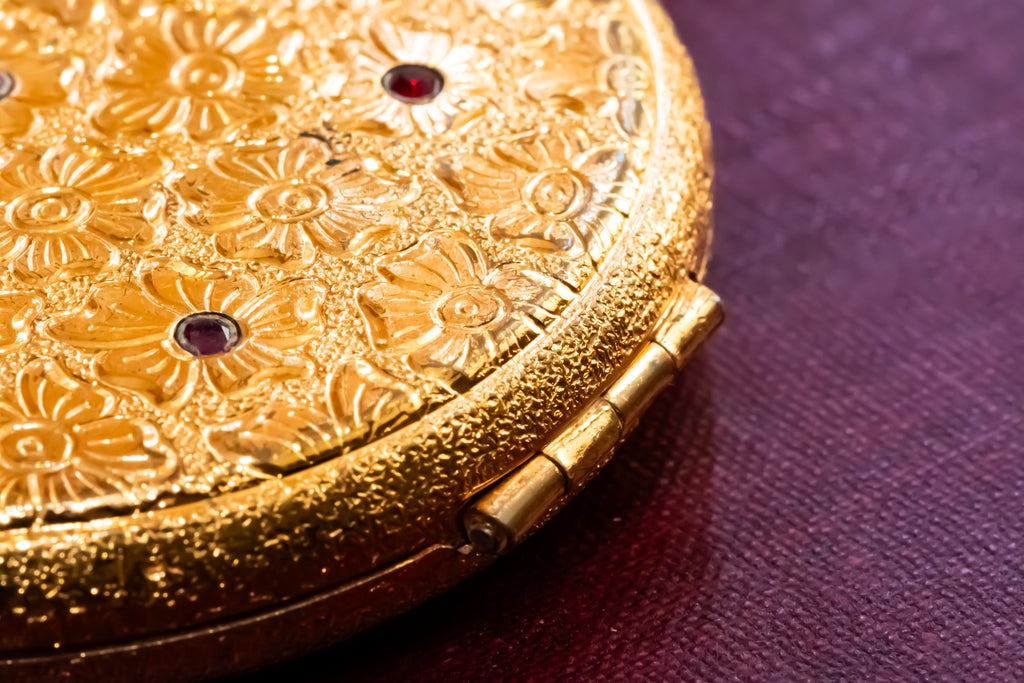 Close-up of an ornate gold compact or case with embossed floral detailing and small dark red gem accents, resting on a textured deep purple surface.