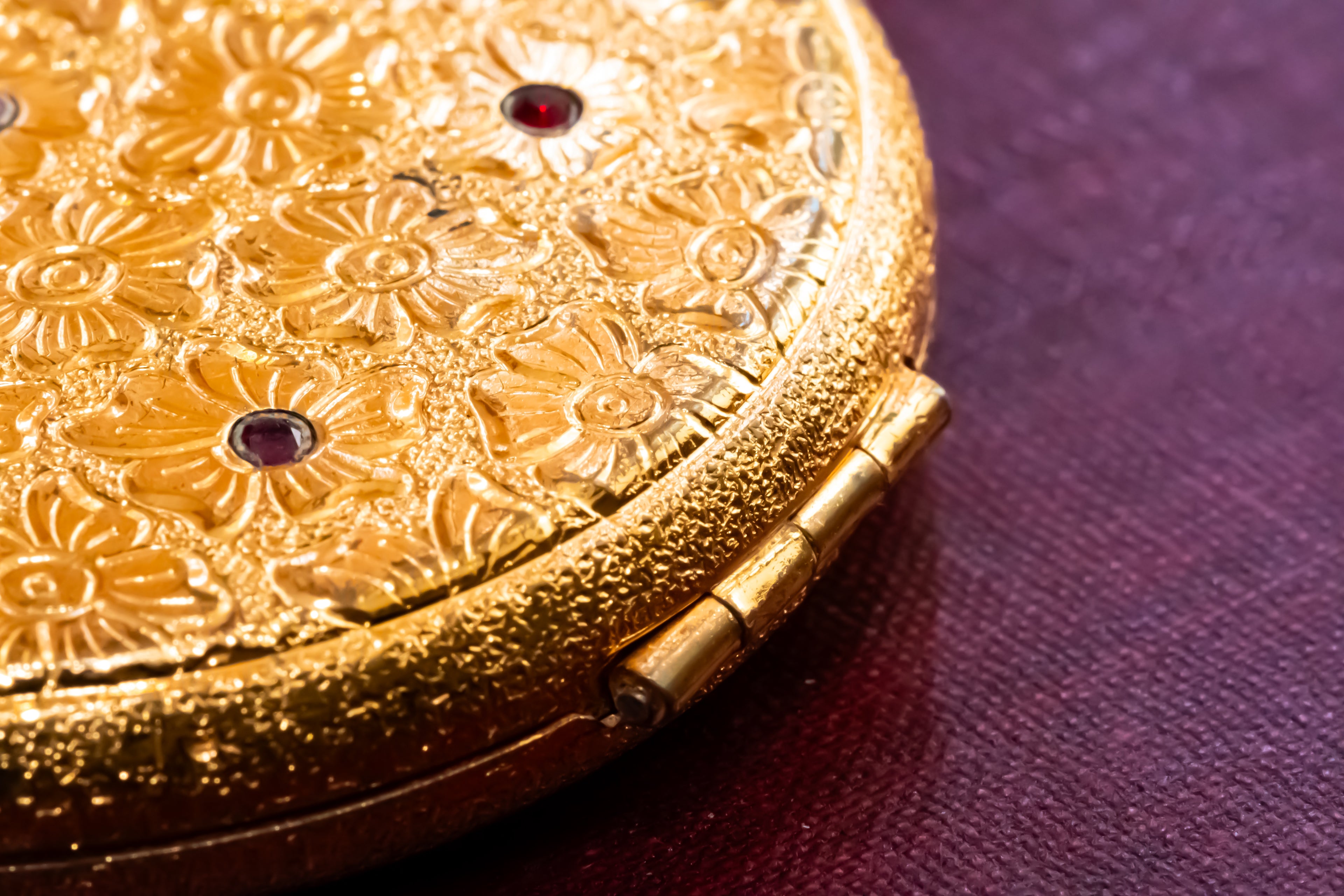 Close-up of an ornate gold compact or case with embossed floral detailing and small dark red gem accents, resting on a textured deep purple surface.