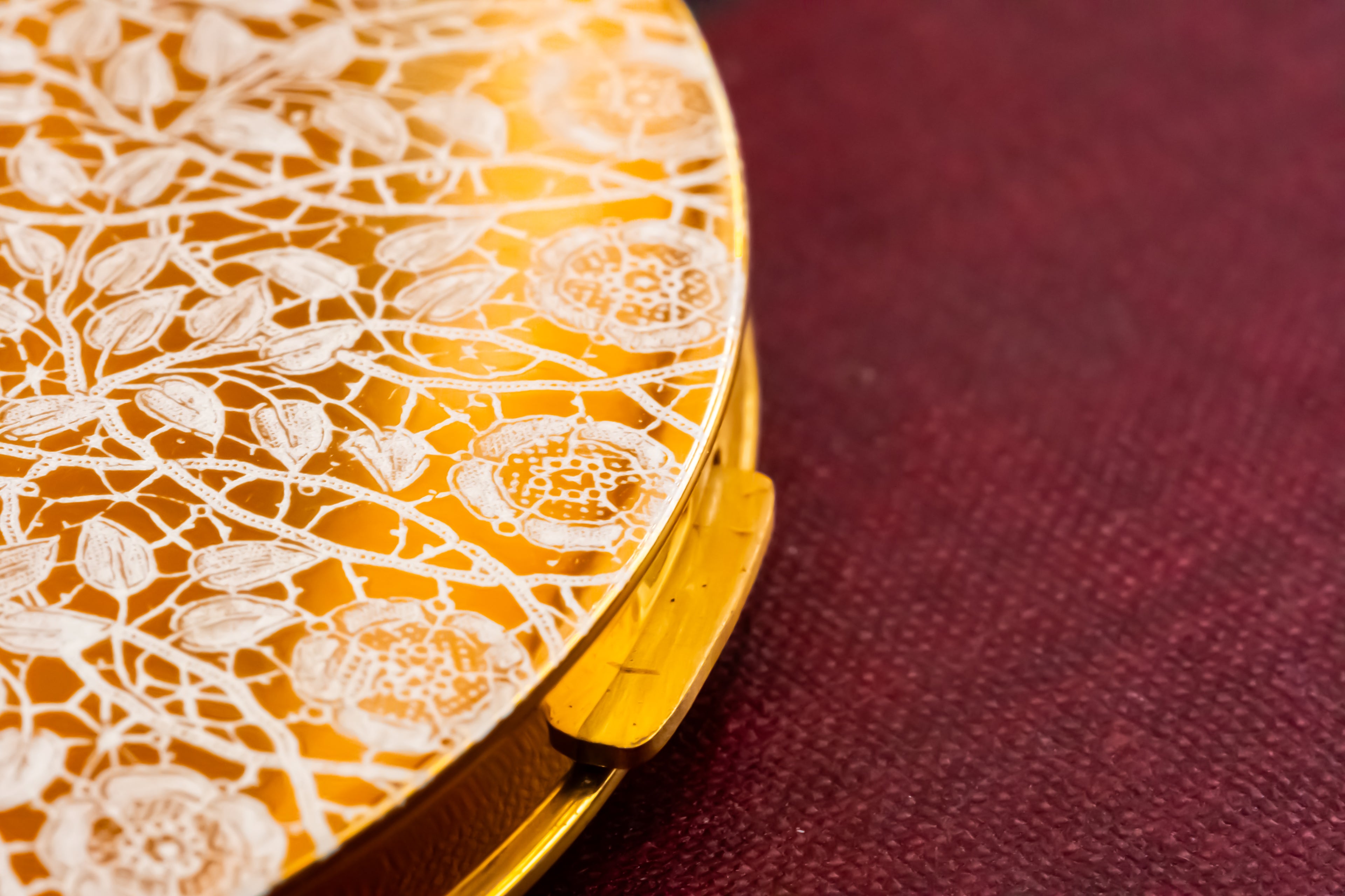 A close-up of a round gold compact or case with an intricate white floral lace-like pattern on the lid, resting on a textured dark burgundy surface.