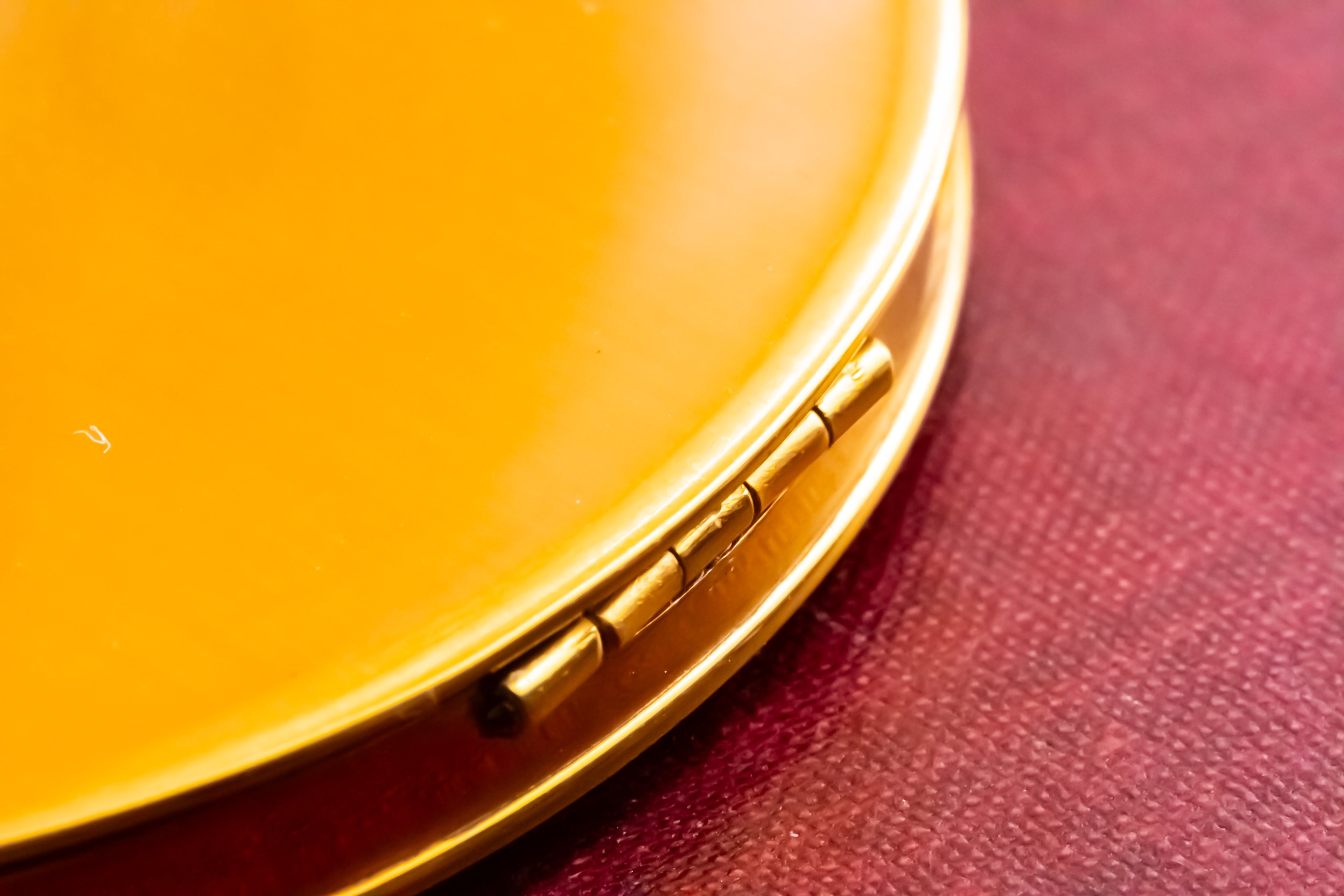 Close-up of a round gold compact case with a small metal hinge, resting on a textured dark red surface.
