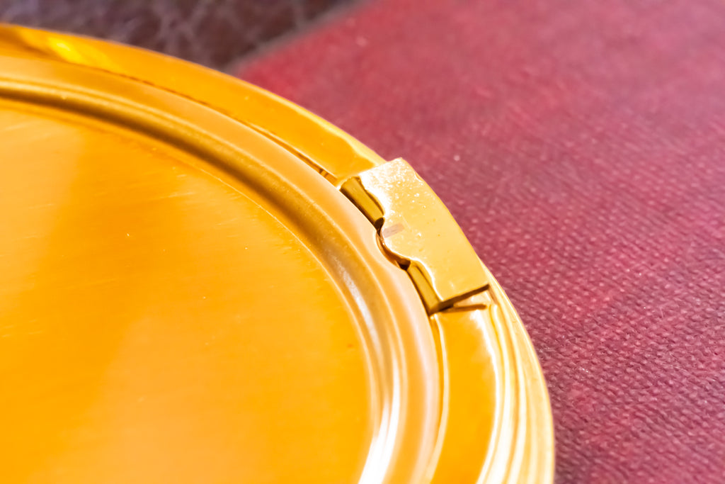 Close-up of a glossy amber-yellow oval powder compact lid with a small metal latch, resting on a textured dark red surface.