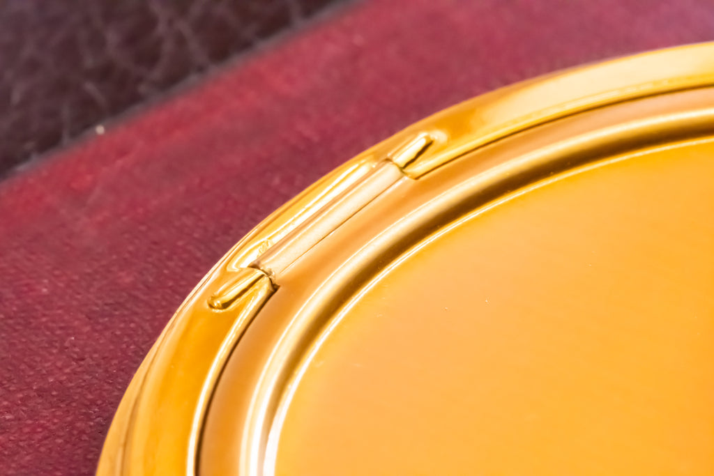 Close-up of a round gold-toned compact mirror or cosmetic case resting on a textured dark red surface.