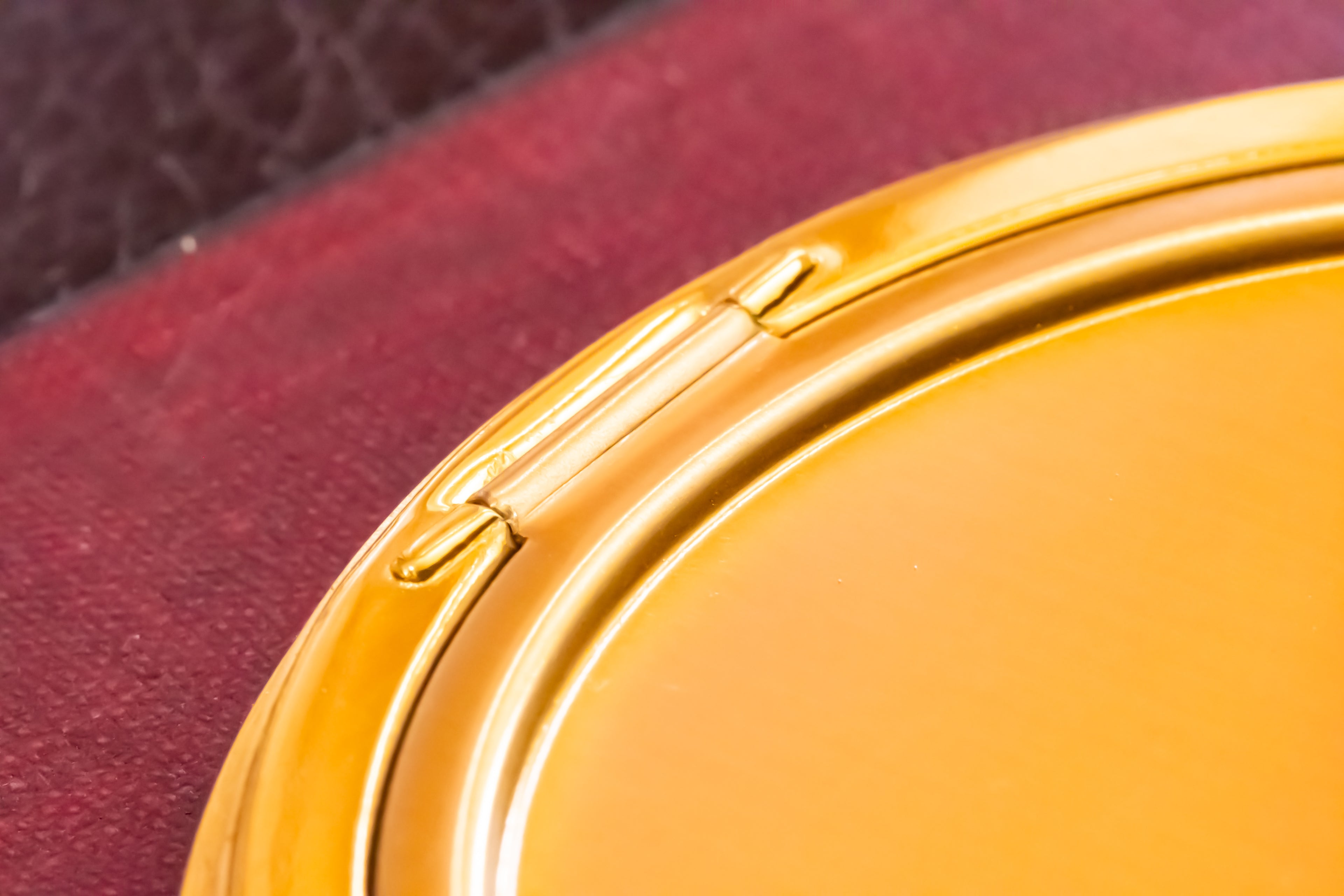 Close-up of a round gold-toned compact mirror or cosmetic case resting on a textured dark red surface.