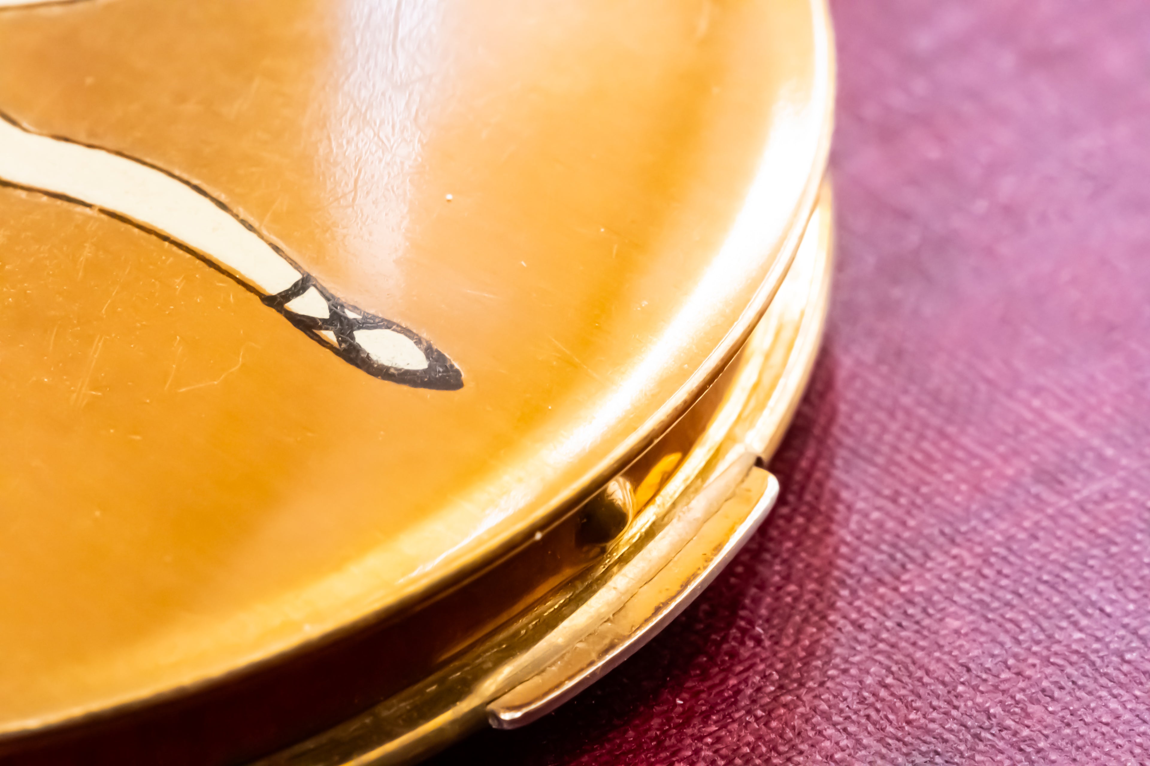 Close-up of a round gold compact case with a small black and white inlay design on the lid, resting on a textured dark red surface.