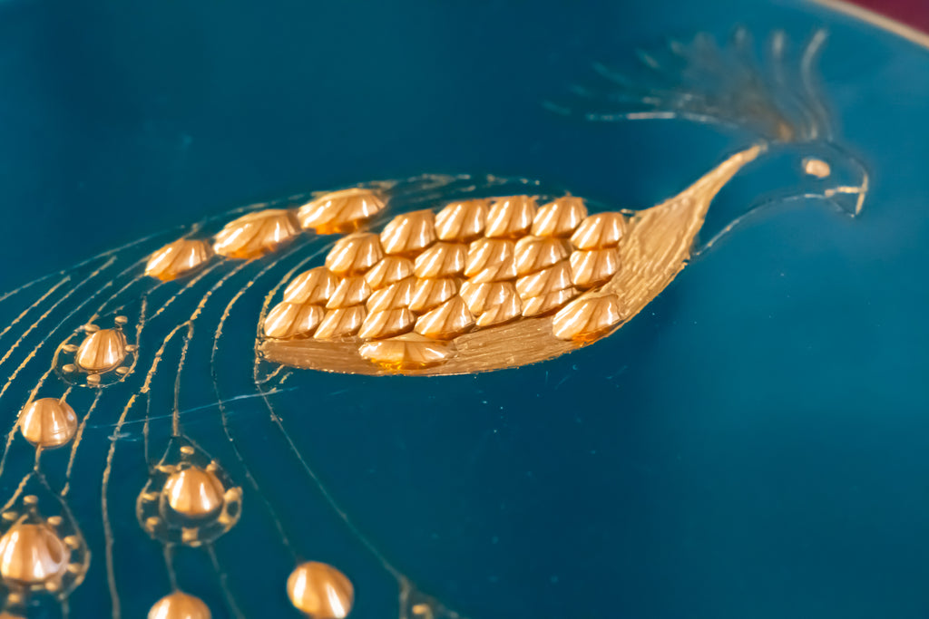 A close-up of a gold peacock motif with detailed feather scales on a deep teal blue background.