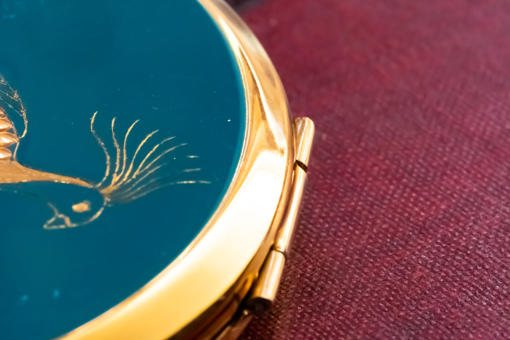 Close-up of a round gold-toned compact or case with a deep teal-blue enamel face and a bird motif, resting on a textured burgundy surface.