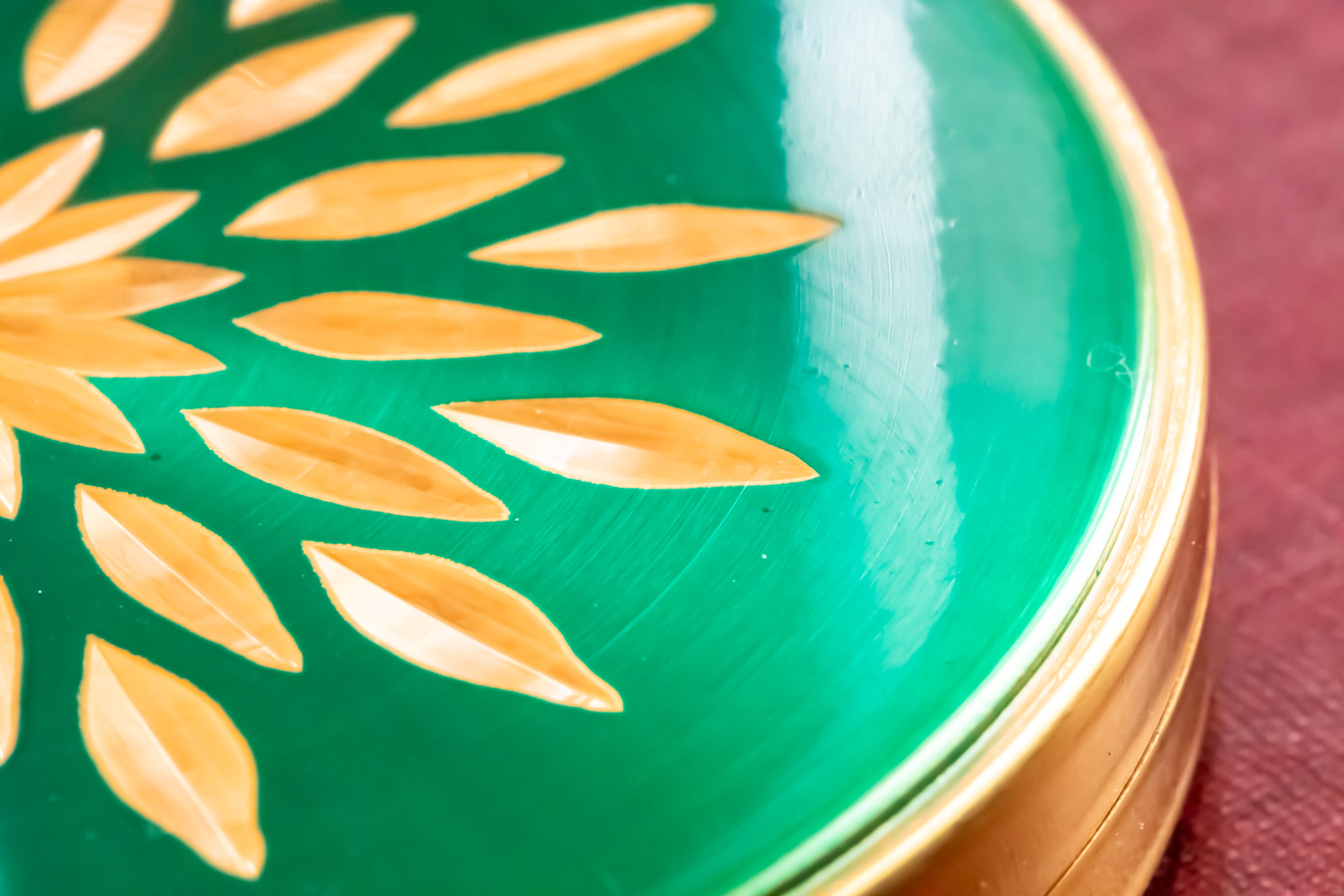 Close-up of a glossy green decorative lid with a gold-toned circular rim and a pattern of gold leaf-like shapes, resting on a dark red surface.