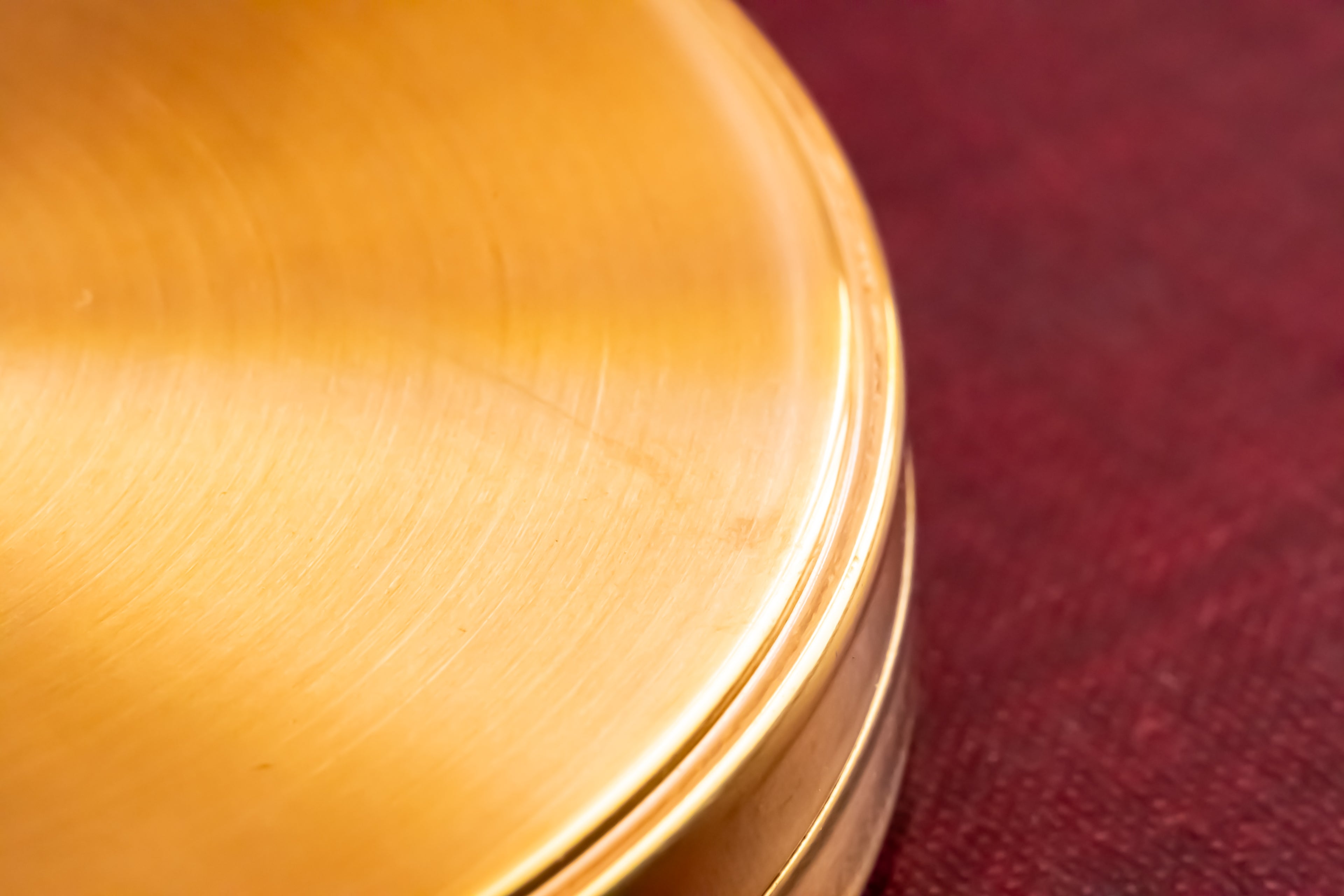 Close-up of a brushed metallic gold circular object, likely a compact case, resting on a textured dark red surface.