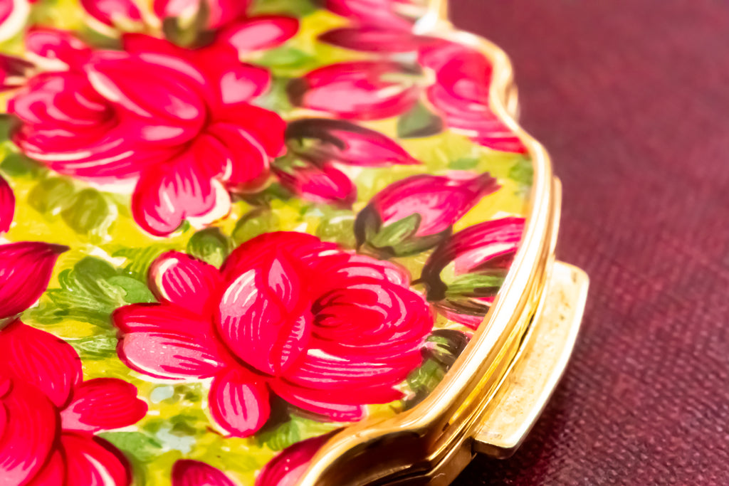A close-up of a decorative compact case featuring a vibrant red rose floral pattern on a yellow-green background, with a gold-toned metal clasp on a textured dark red surface.