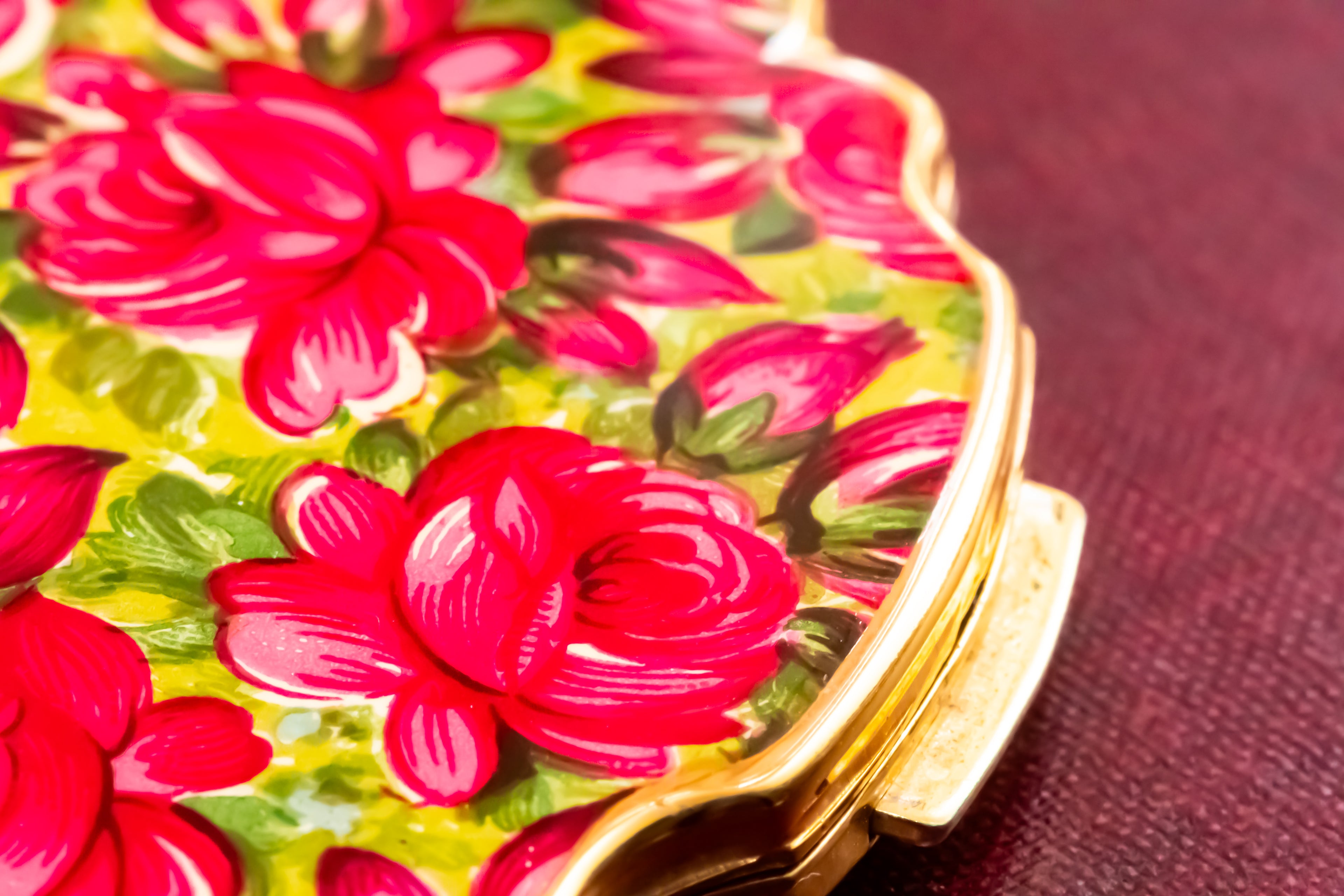 A close-up of a decorative compact case featuring a vibrant red rose floral pattern on a yellow-green background, with a gold-toned metal clasp on a textured dark red surface.