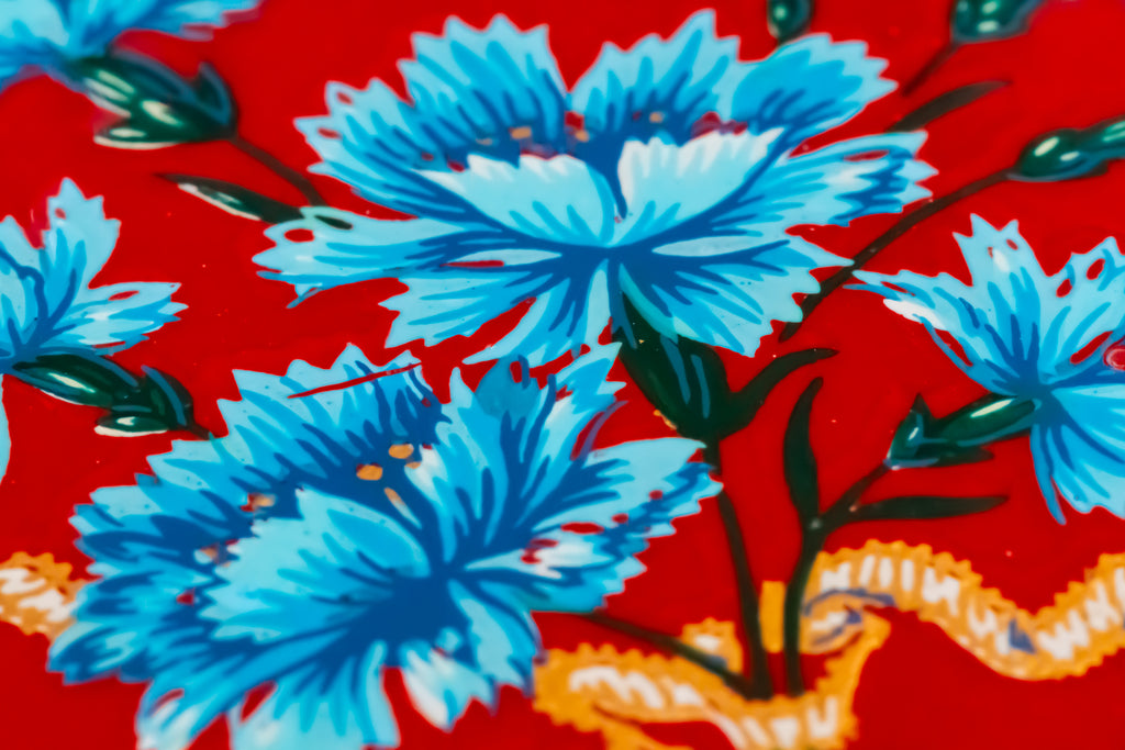 A close-up of a red patterned surface featuring large blue flowers with dark green leaves and stems, plus a small orange decorative element near the bottom.