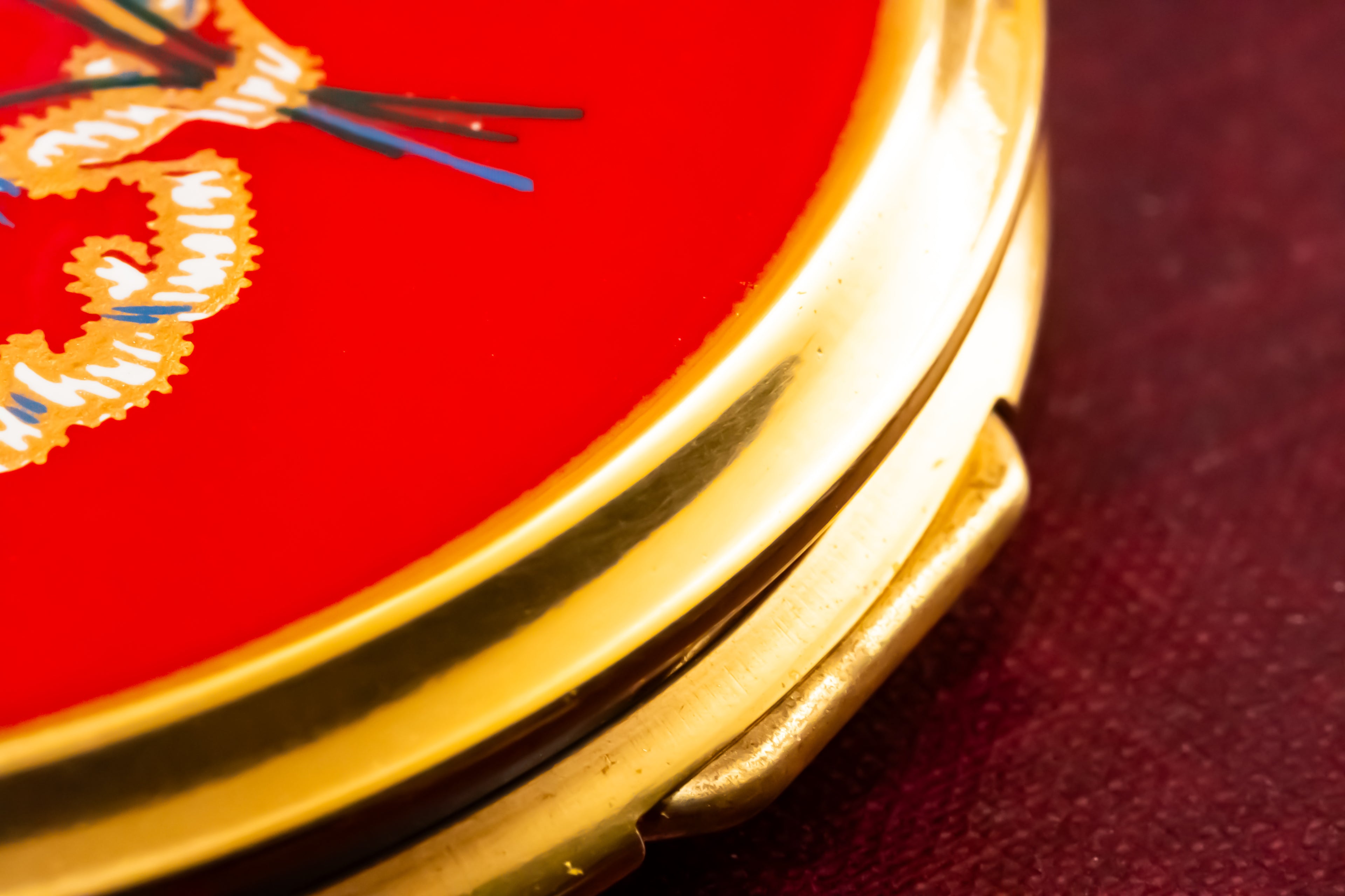Close-up of a round compact mirror with a polished gold-tone metal frame and a bright red velvet cover featuring embroidered gold and blue details.
