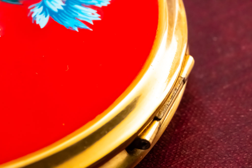 Close-up of a round compact with a bright red lid featuring a blue floral design and a polished gold-tone metal edge, resting on a dark textured surface.