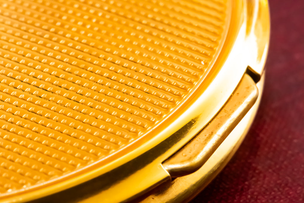 Close-up of a round gold compact case with a textured lid and a small hinge tab, set against a dark red background.