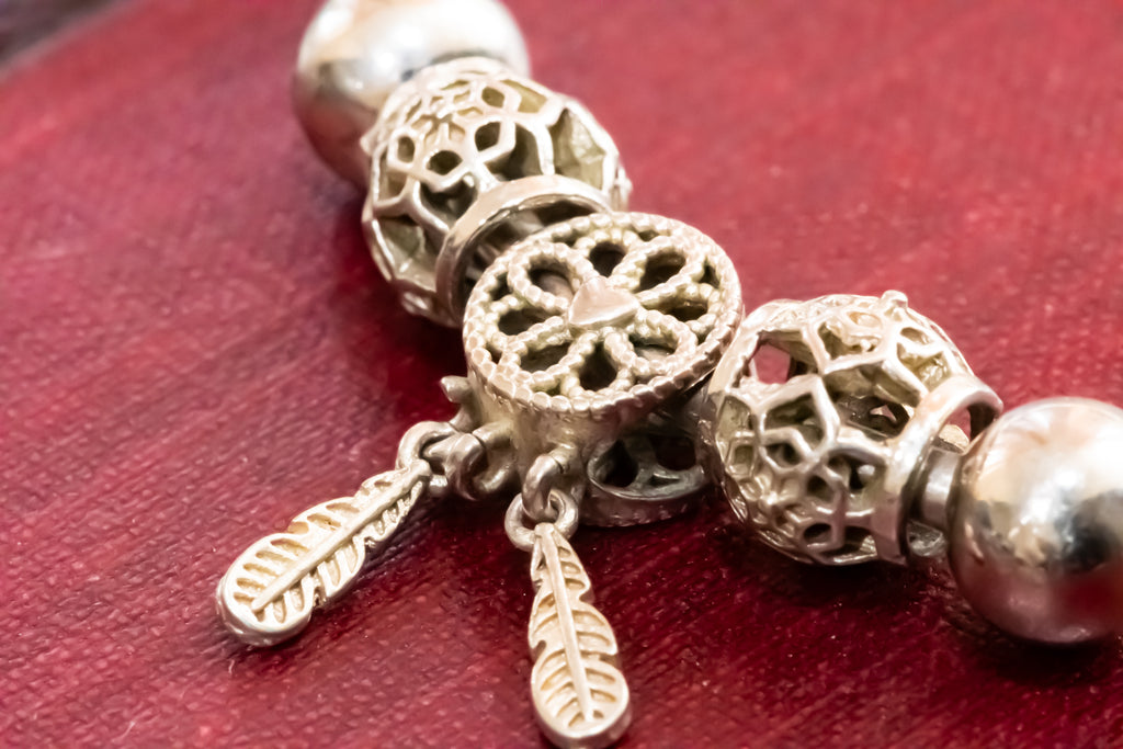 A close-up of a silver-toned beaded bracelet featuring ornate filigree beads, a central circular bead with a floral cutout pattern, and two small feather-shaped charms, displayed on a textured red surface.