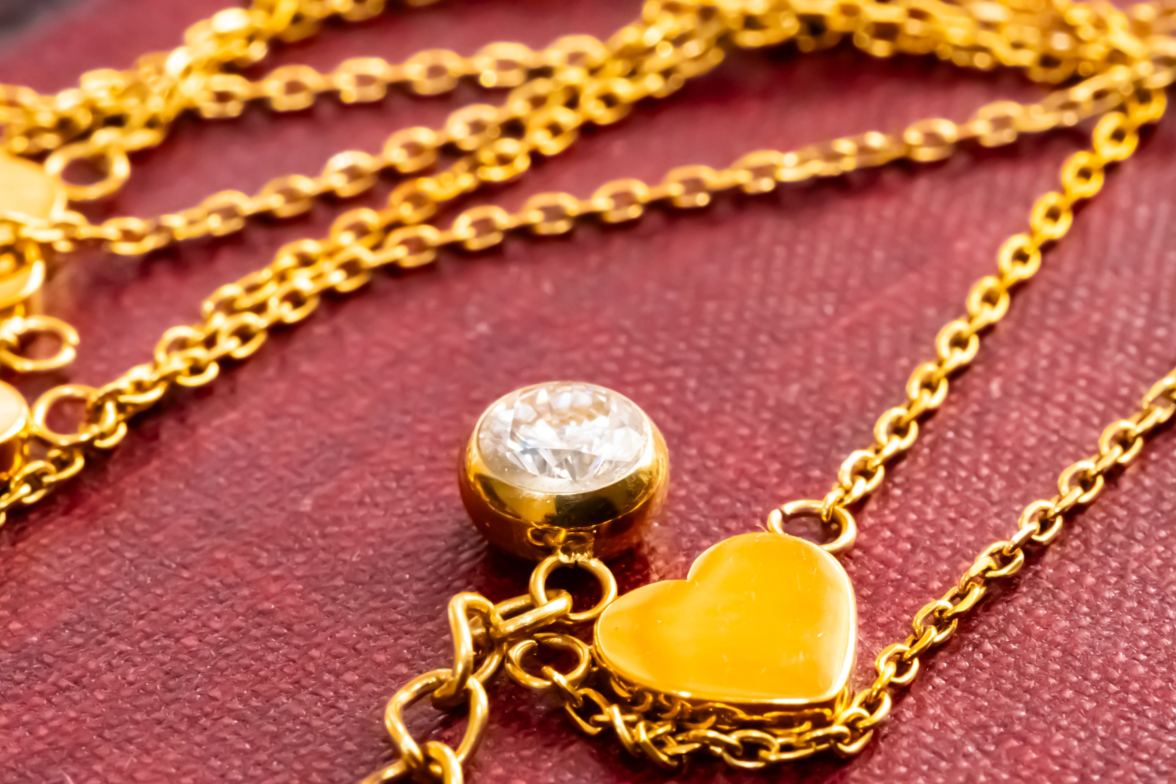Gold chain necklace with a small round clear stone bead and a polished gold heart pendant, displayed on a textured dark red fabric background.