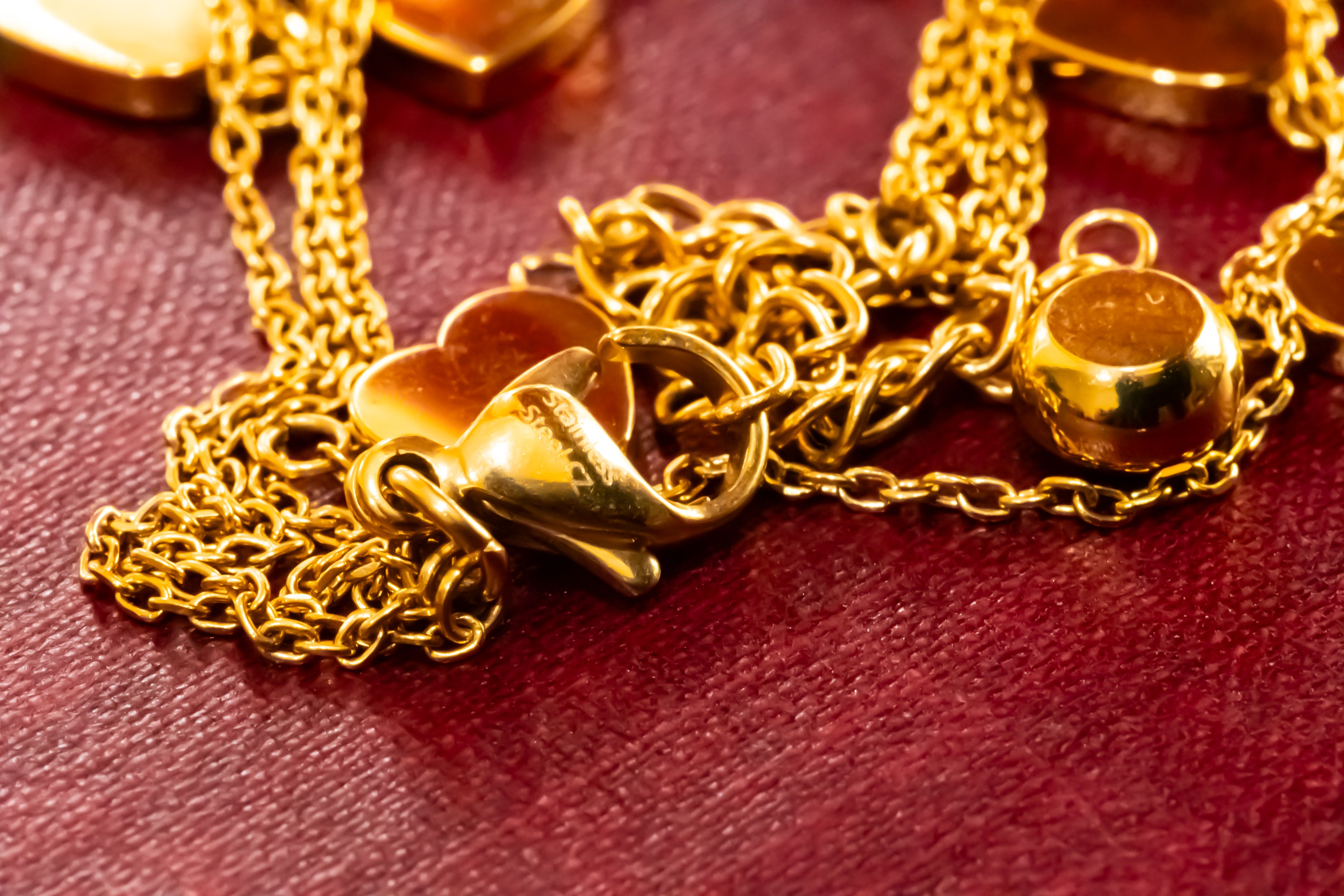 A close-up of a gold-tone chain necklace with heart-shaped charms and a round pendant, resting on a textured dark red surface.