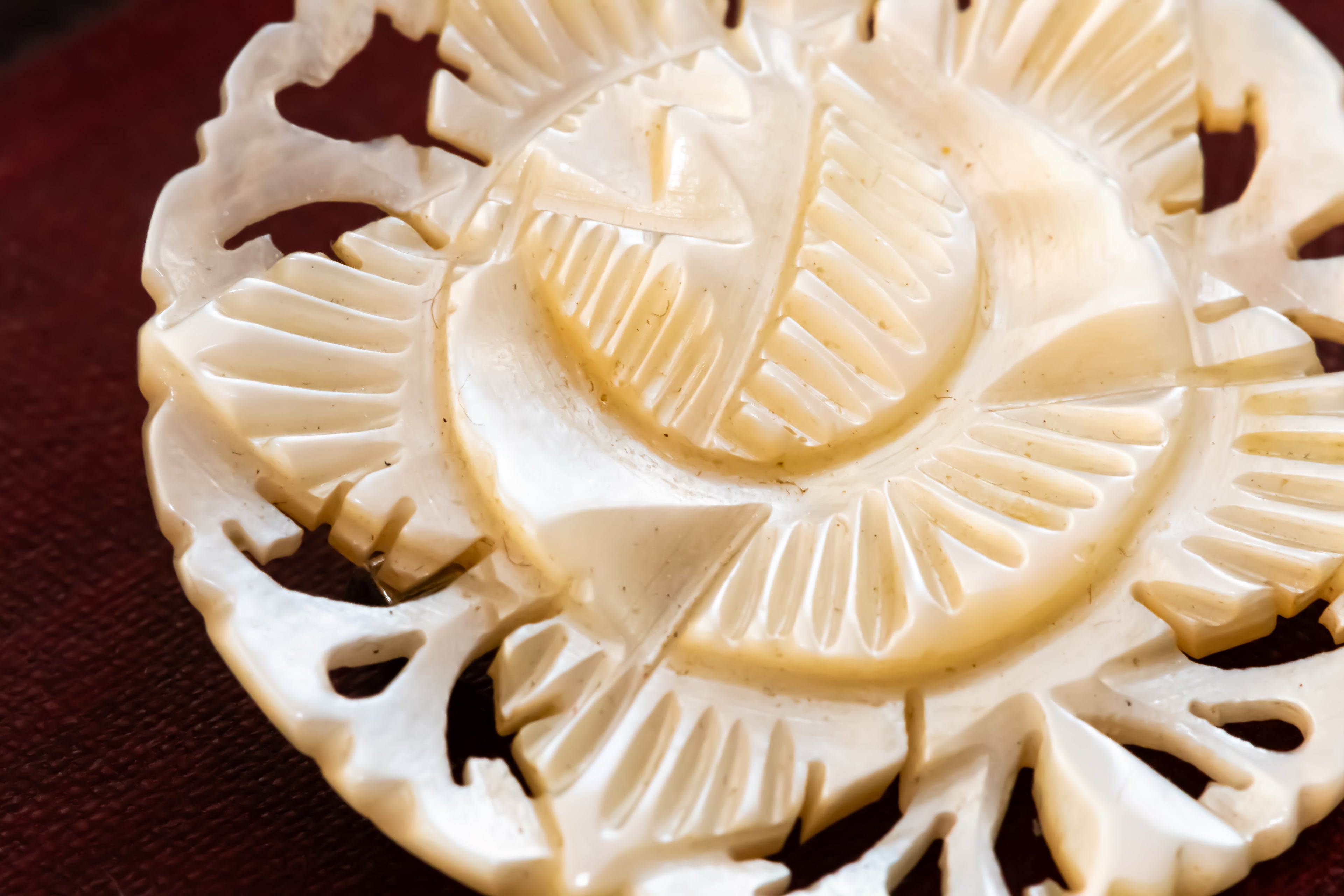 A close-up of a carved circular brooch made from a warm mother of pearl material, featuring a sunburst or floral cutout design, resting on a dark red textured surface.