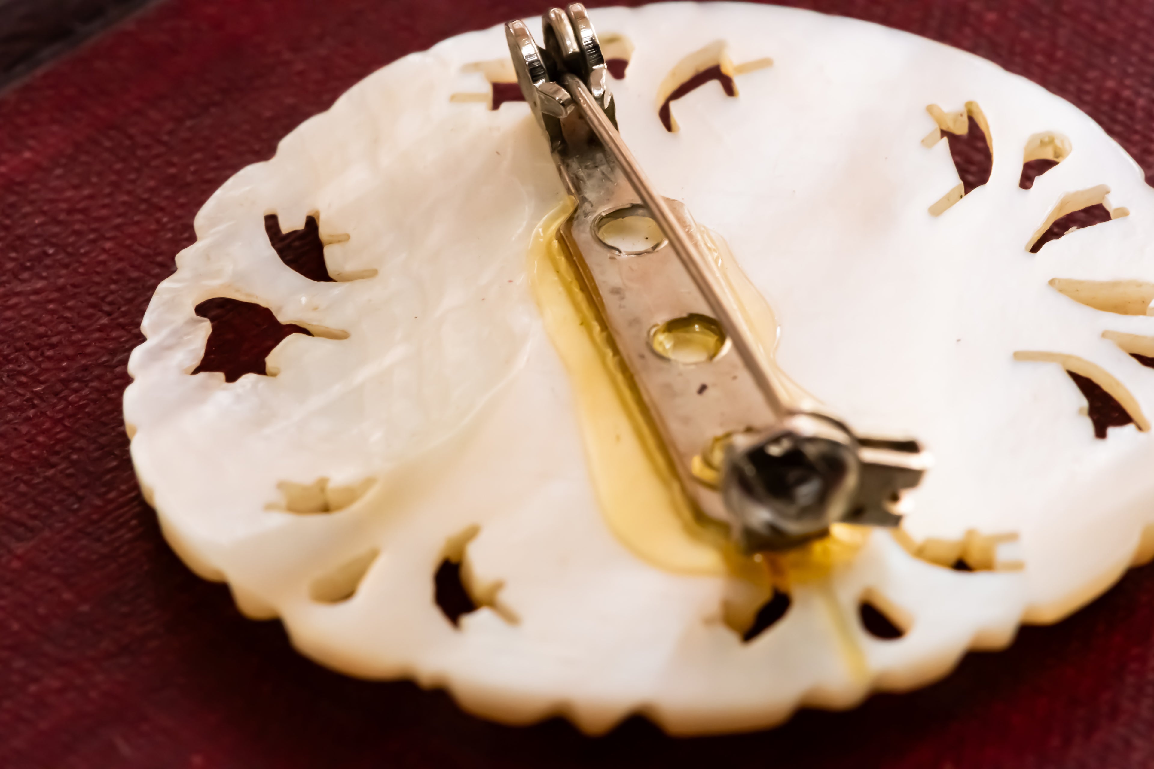 A close-up of a circular white mother of pearl brooch with intricate cutout details and a silver safety pin pinback, resting on a dark red textured surface.