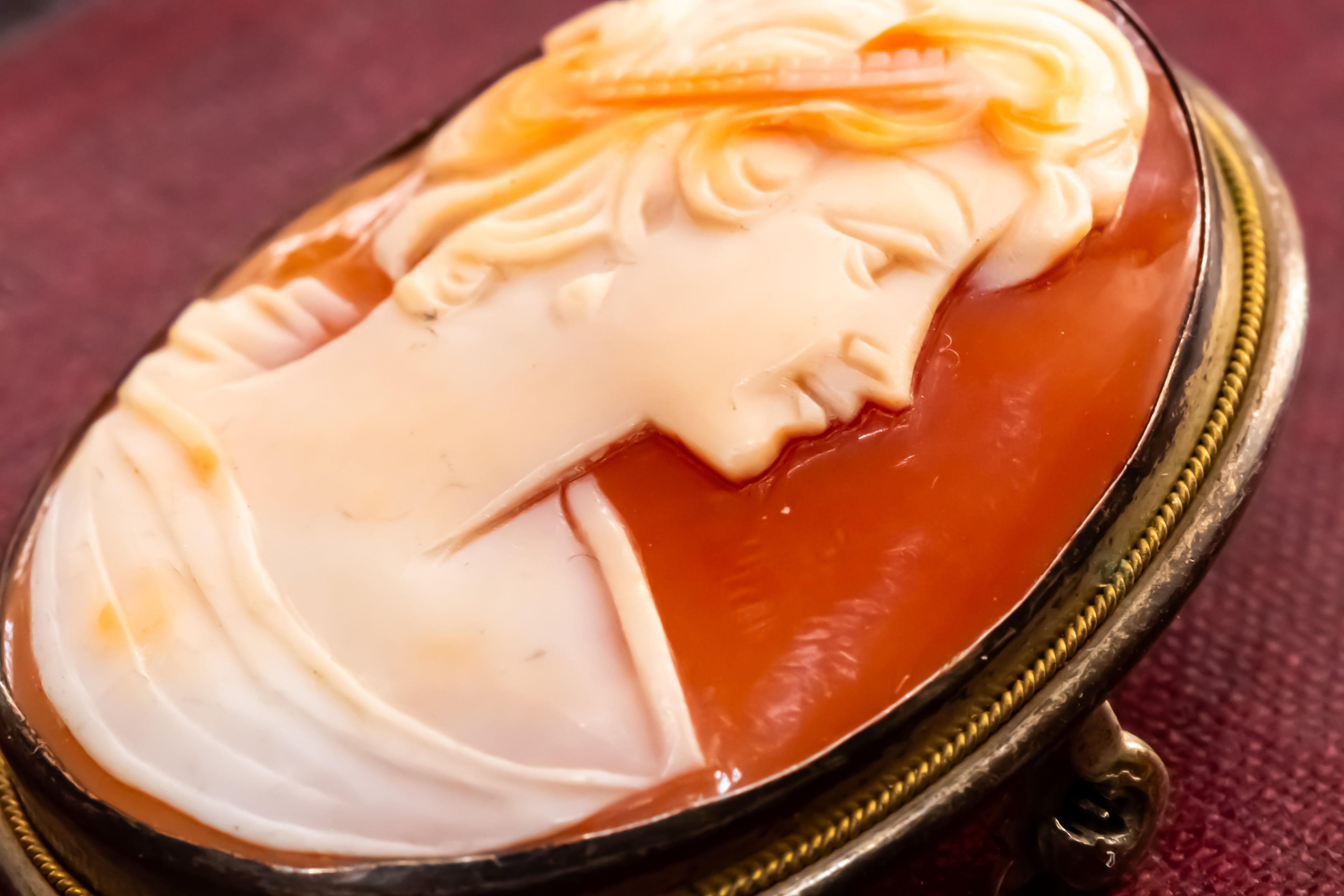An oval carved cameo-style pendant or brooch featuring a profile portrait in warm ivory and peach tones over a translucent amber-orange background, set in a dark metallic frame with a gold-toned inner border.