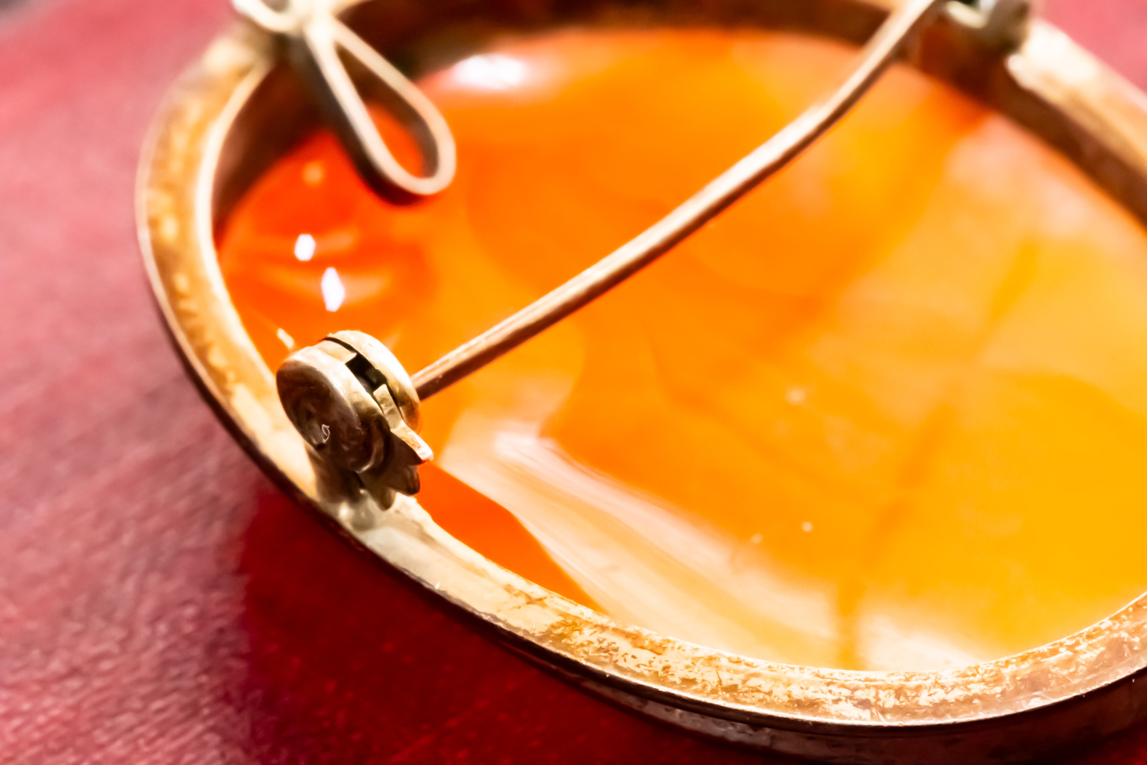 Close-up of a round amber-orange shell back with a gold-toned metal frame and a small looped metal piece resting on top, set against a dark red background.