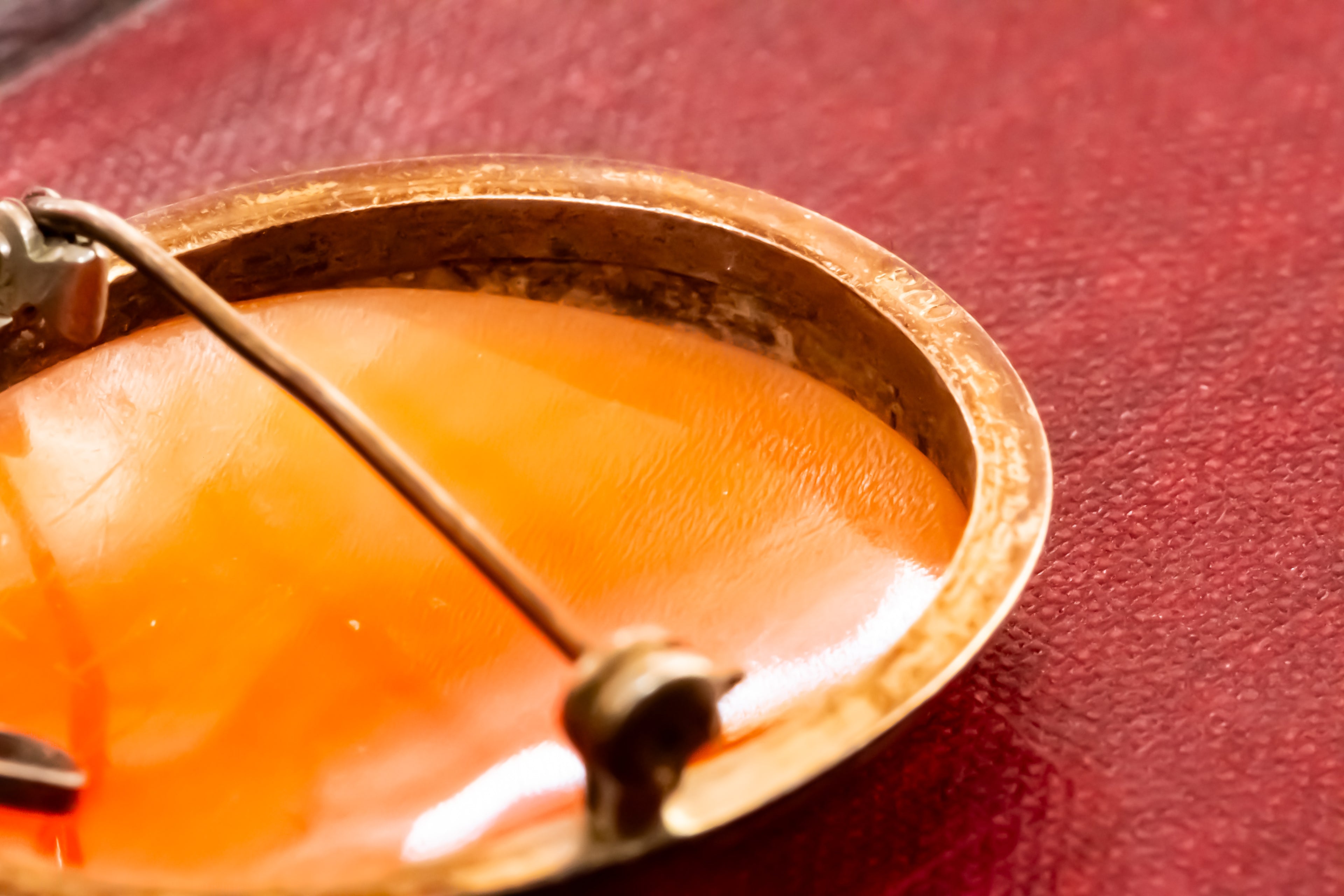 Close-up of a round vintage brooch with a warm orange shell center and a gold-toned metal rim, resting on a textured red surface.