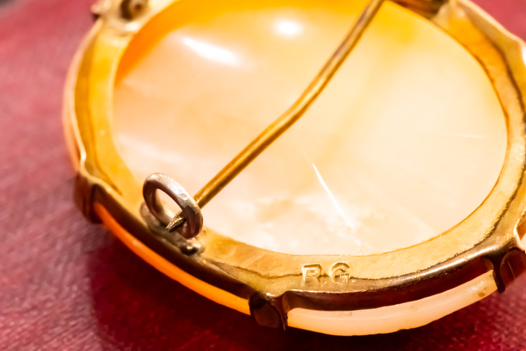 Close-up of the back of a round gold-tone brooch or pendant with a pale cream inlay and a pin fastener visible, resting on a dark red surface.