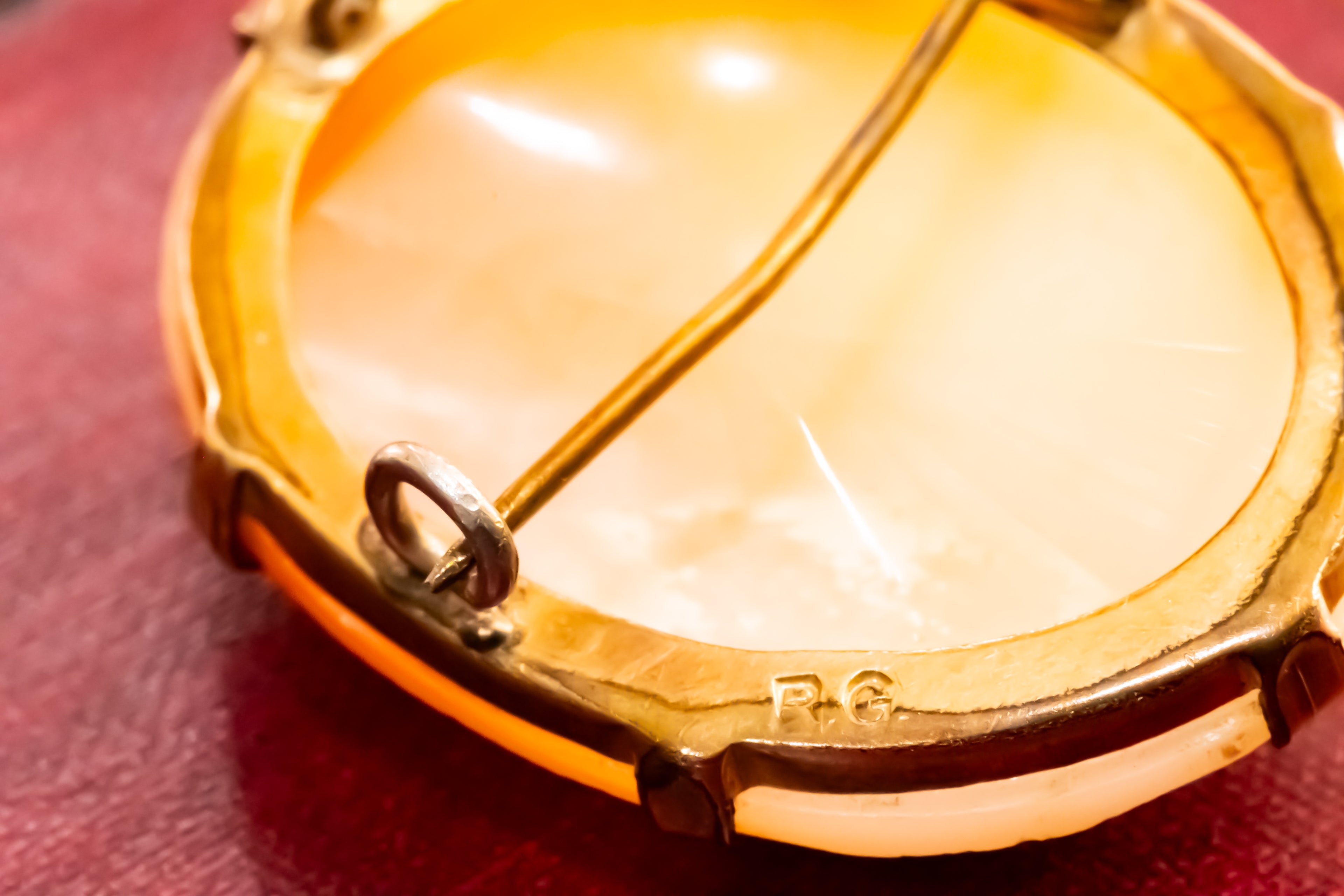 Close-up of the back of a round gold-tone brooch or pendant with a pale cream inlay and a pin fastener visible, resting on a dark red surface.